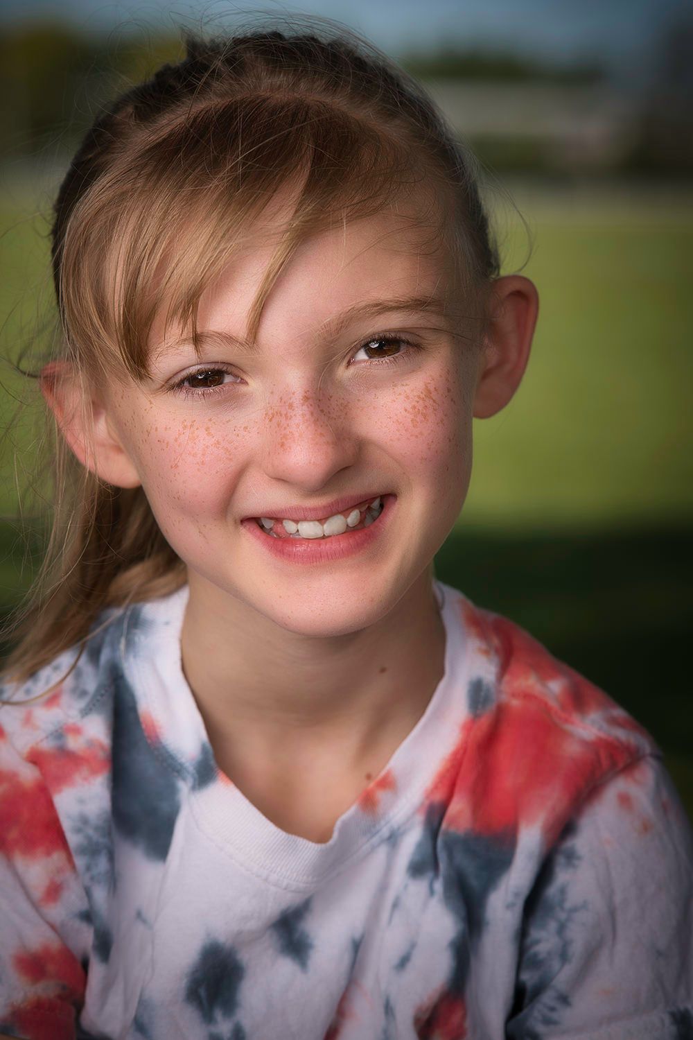 A cheerful freckled girl with red hair smiles brightly wearing a tie-dye shirt in an outdoor setting.