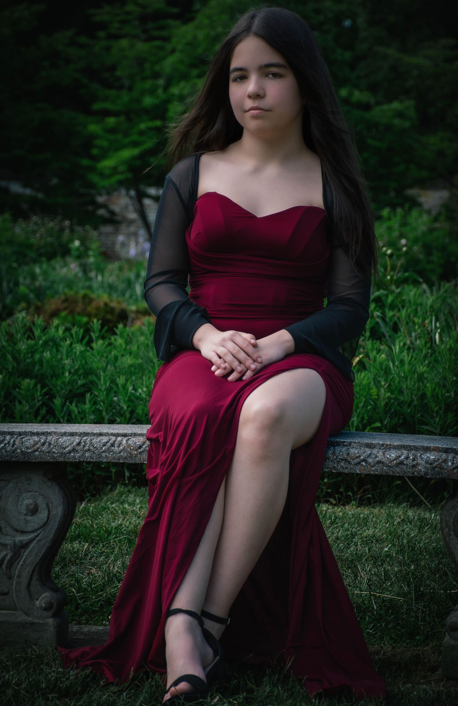 Young woman in elegant burgundy strapless gown with black sheer wrap seated on stone bench in garden.