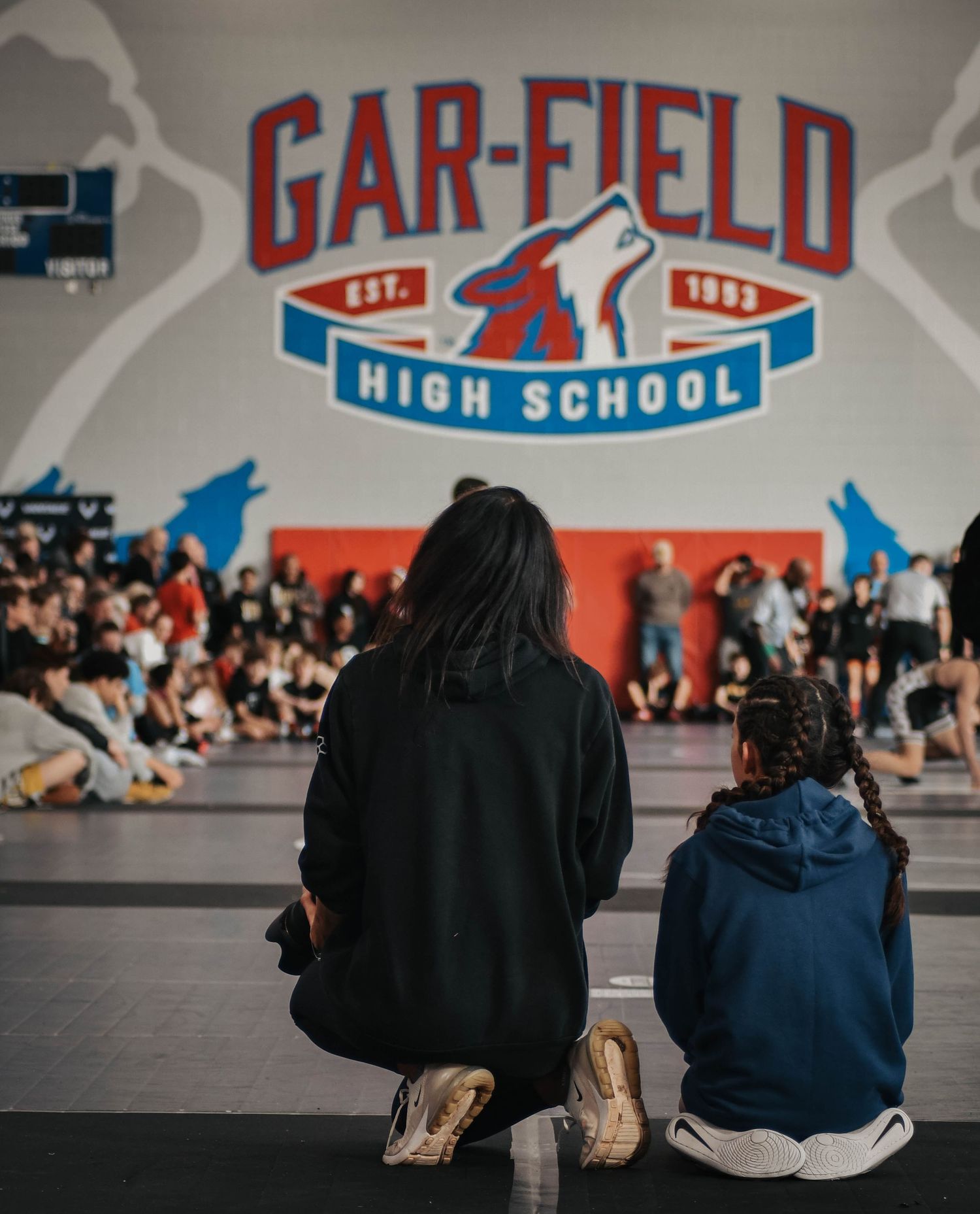 Two spectators kneeling courtside watching wrestling tournament at Gar-Field High School gym.