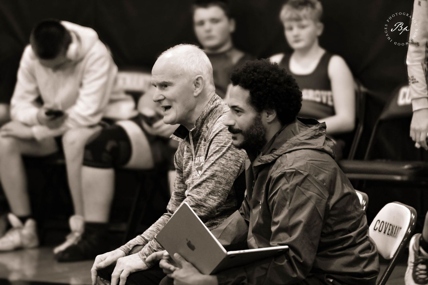 Black and white photo of two wrestling coaches reviewing notes together on a laptop at a meet.