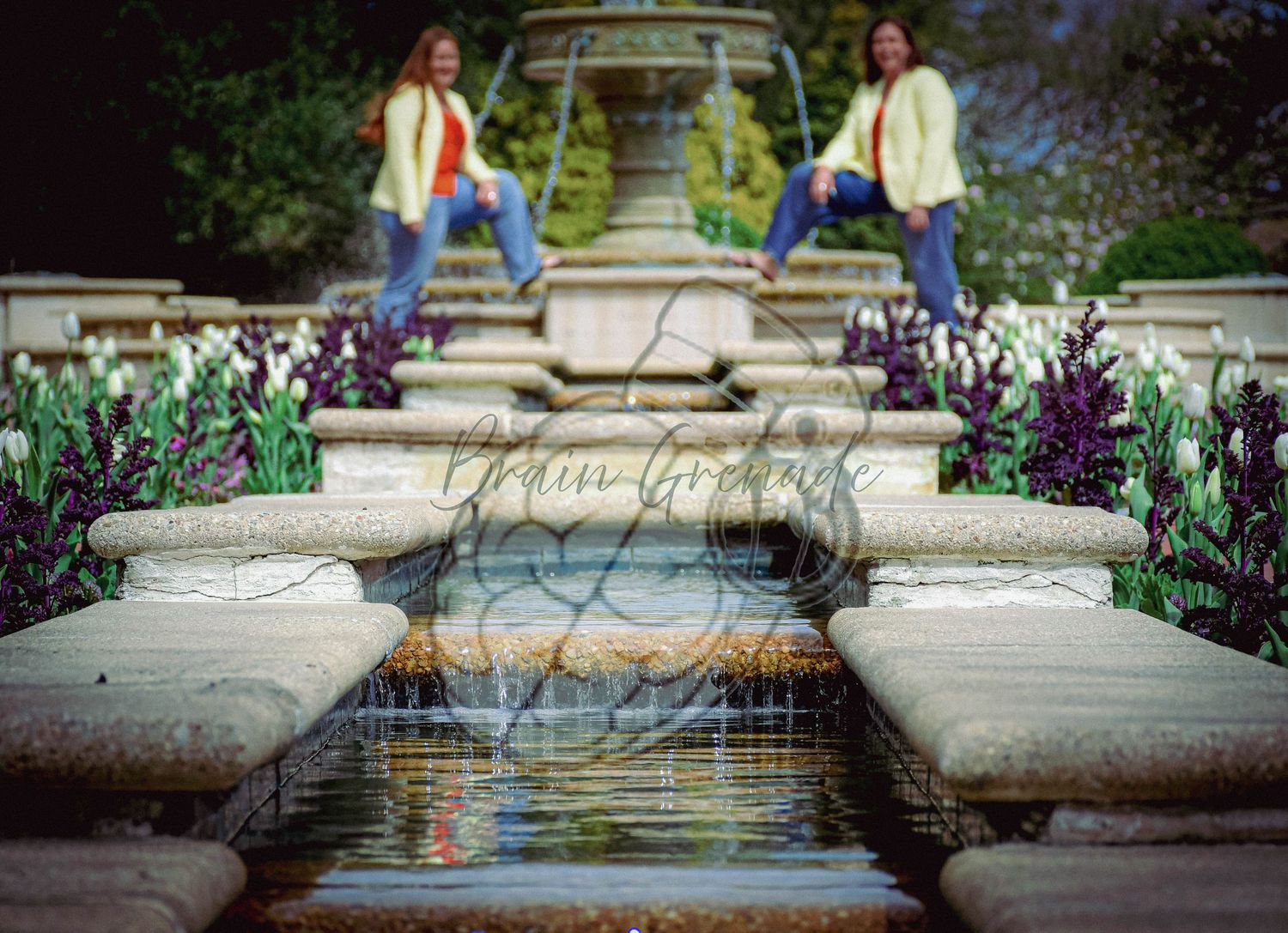 Two women in yellow blazers pose at ornate fountain surrounded by tulips in garden.