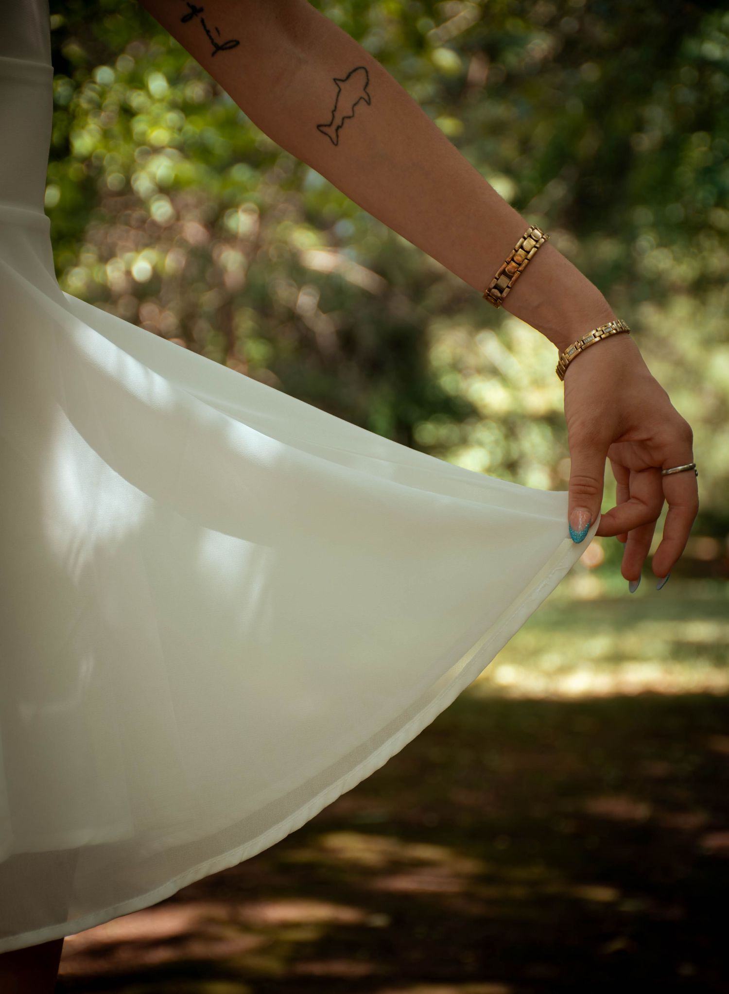 Woman in white dress holding skirt hem, showing gold bracelet, ring, blue nails, and arm tattoos outdoors.