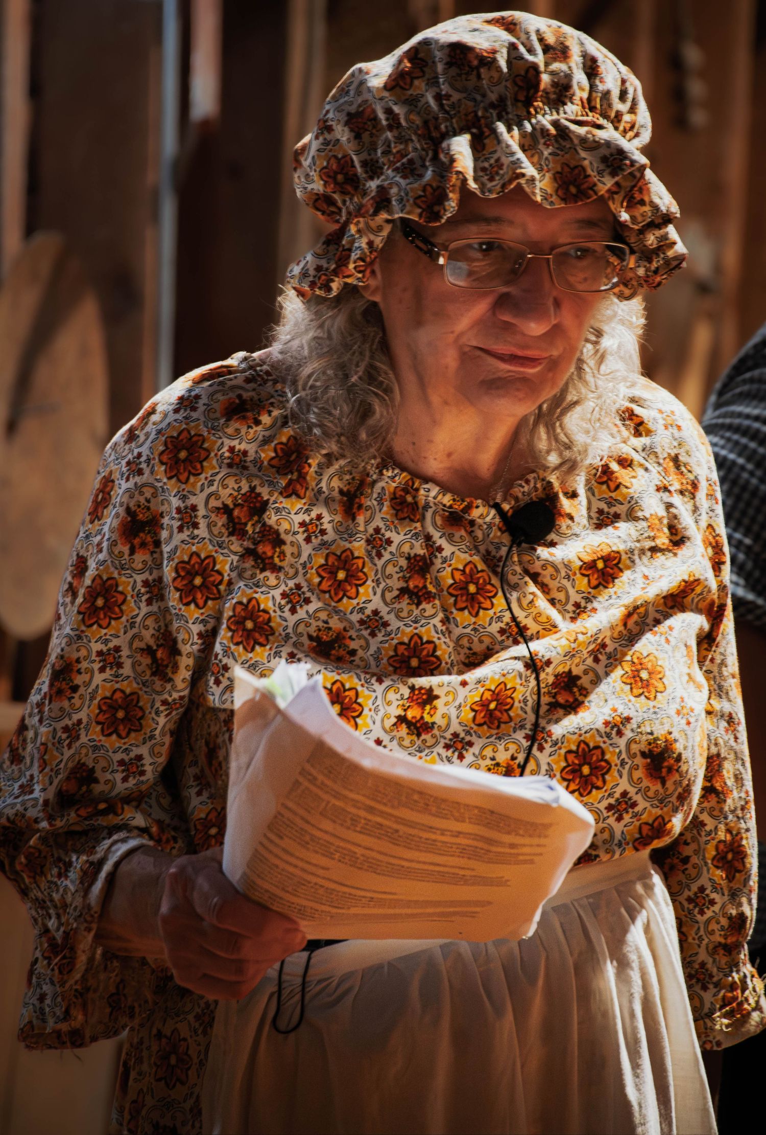Elderly woman in vintage floral colonial costume and bonnet holding papers at a historical reenactment event.