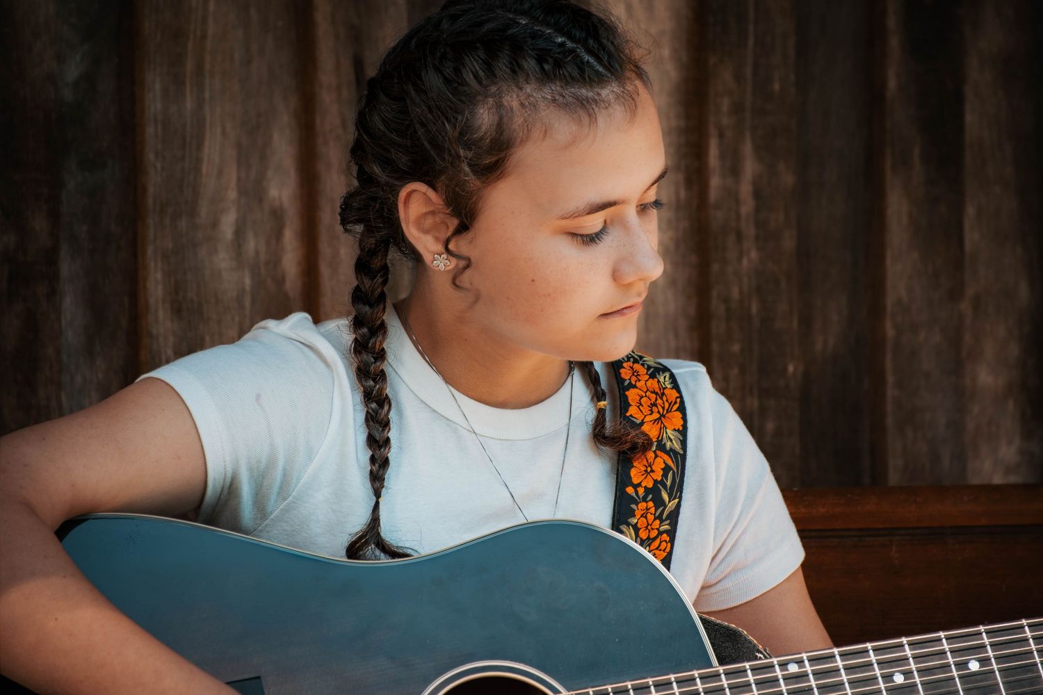Young girl with braided hair playing acoustic guitar with floral strap, wearing white t-shirt outdoors.