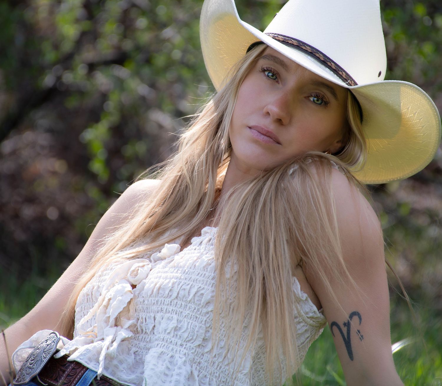 Blonde woman in white cowboy hat and lace top with belt sitting outdoors in green nature setting.
