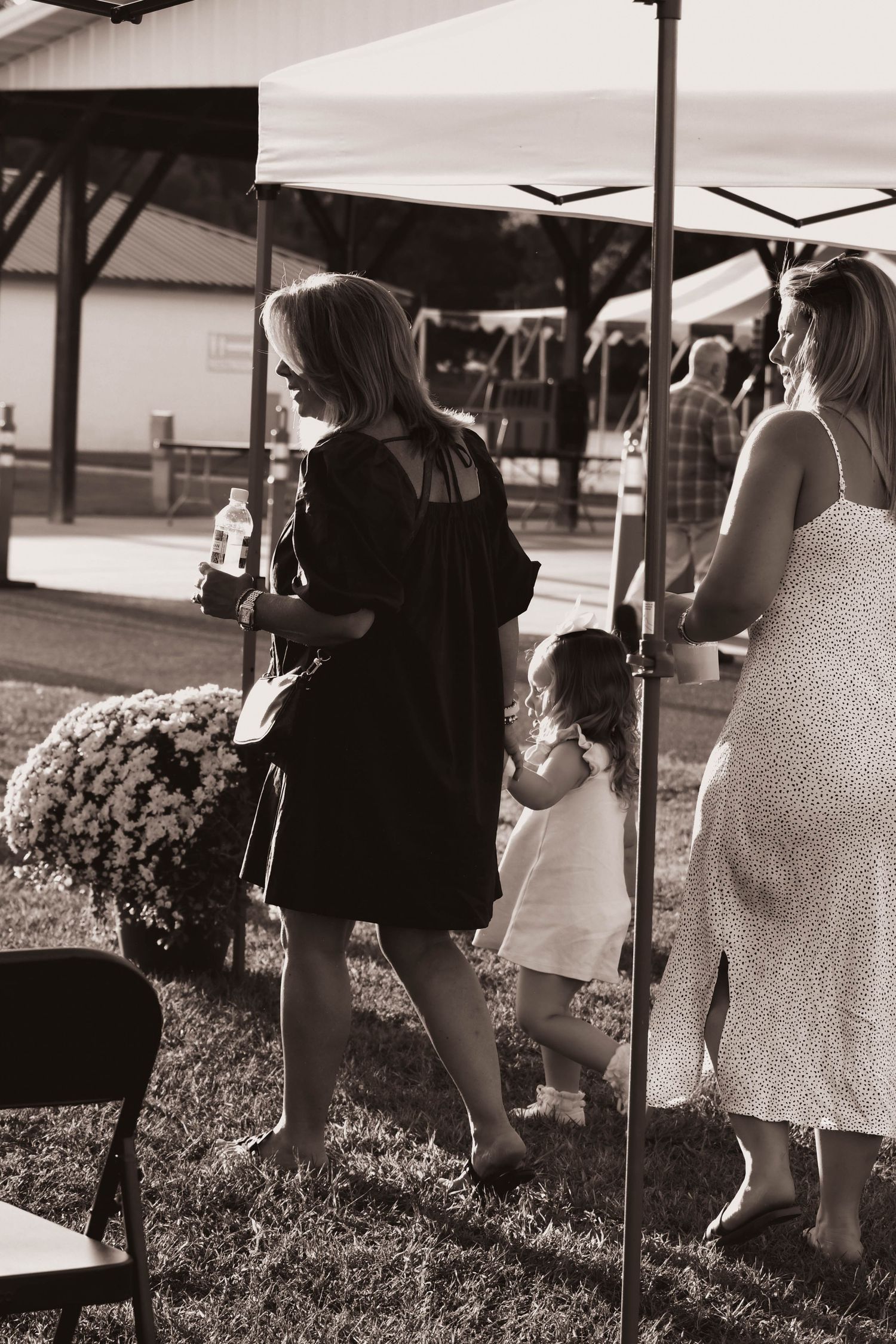 Black and white photo of women and a toddler walking under a tent at an outdoor event.