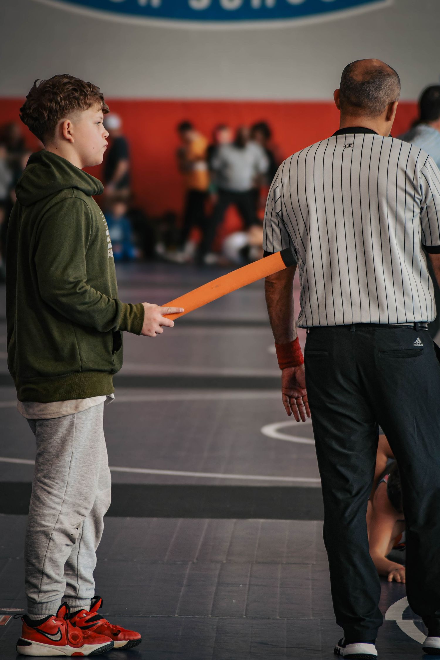 Young boy holding orange card stands beside a wrestling referee in striped shirt during a match.