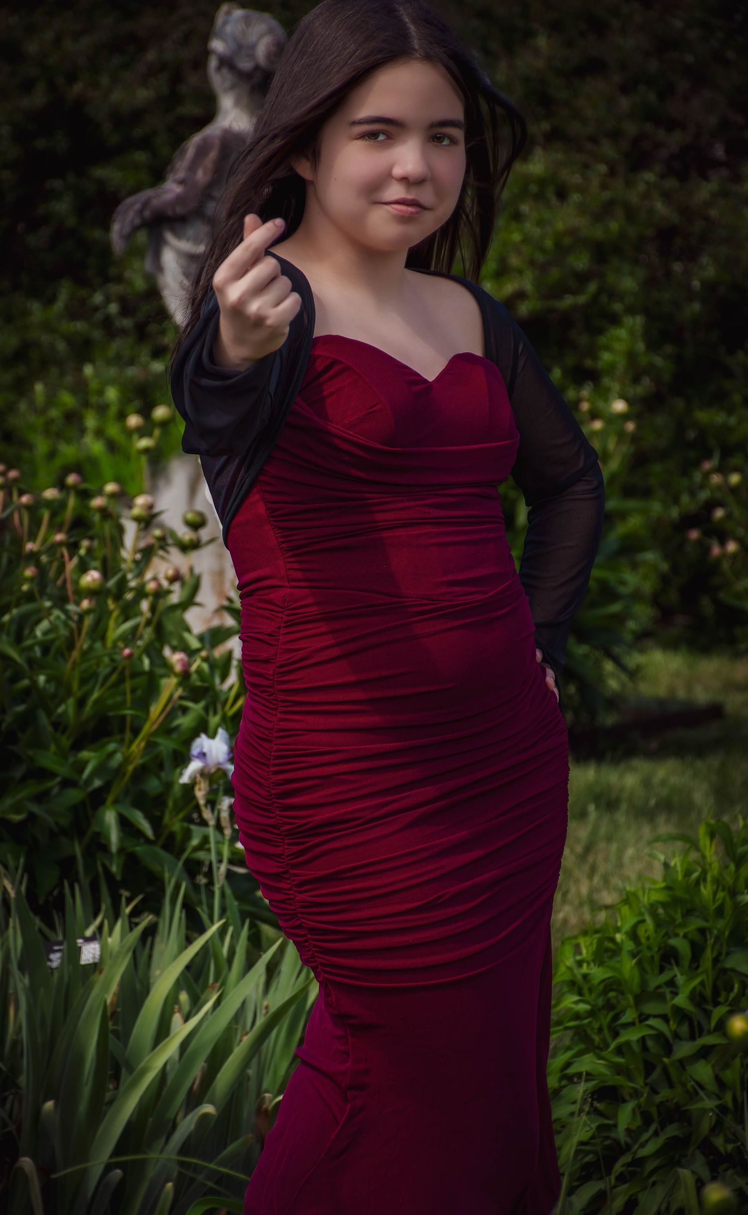 Young woman in red ruched strapless dress with black sheer wrap posing among garden flowers.