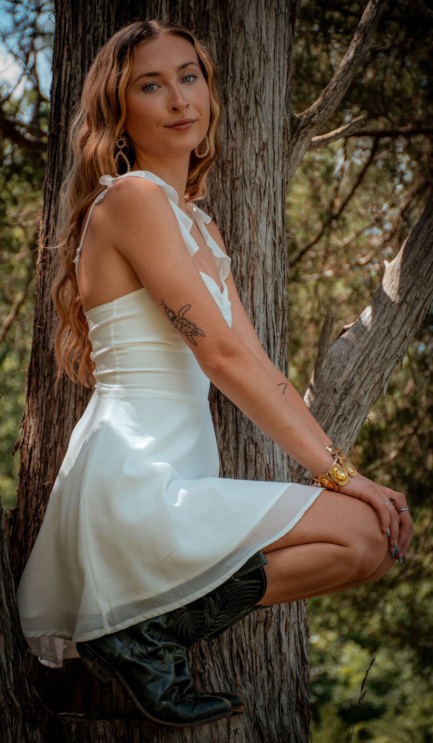 Woman in white dress and cowboy boots posing against a tree outdoors.