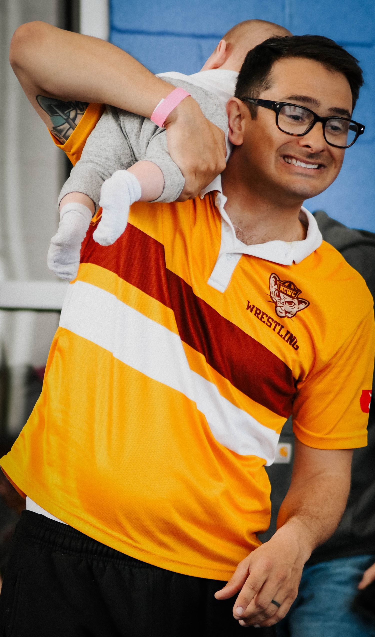 Smiling man in yellow wrestling shirt holds a baby on his shoulder at a wrestling event.