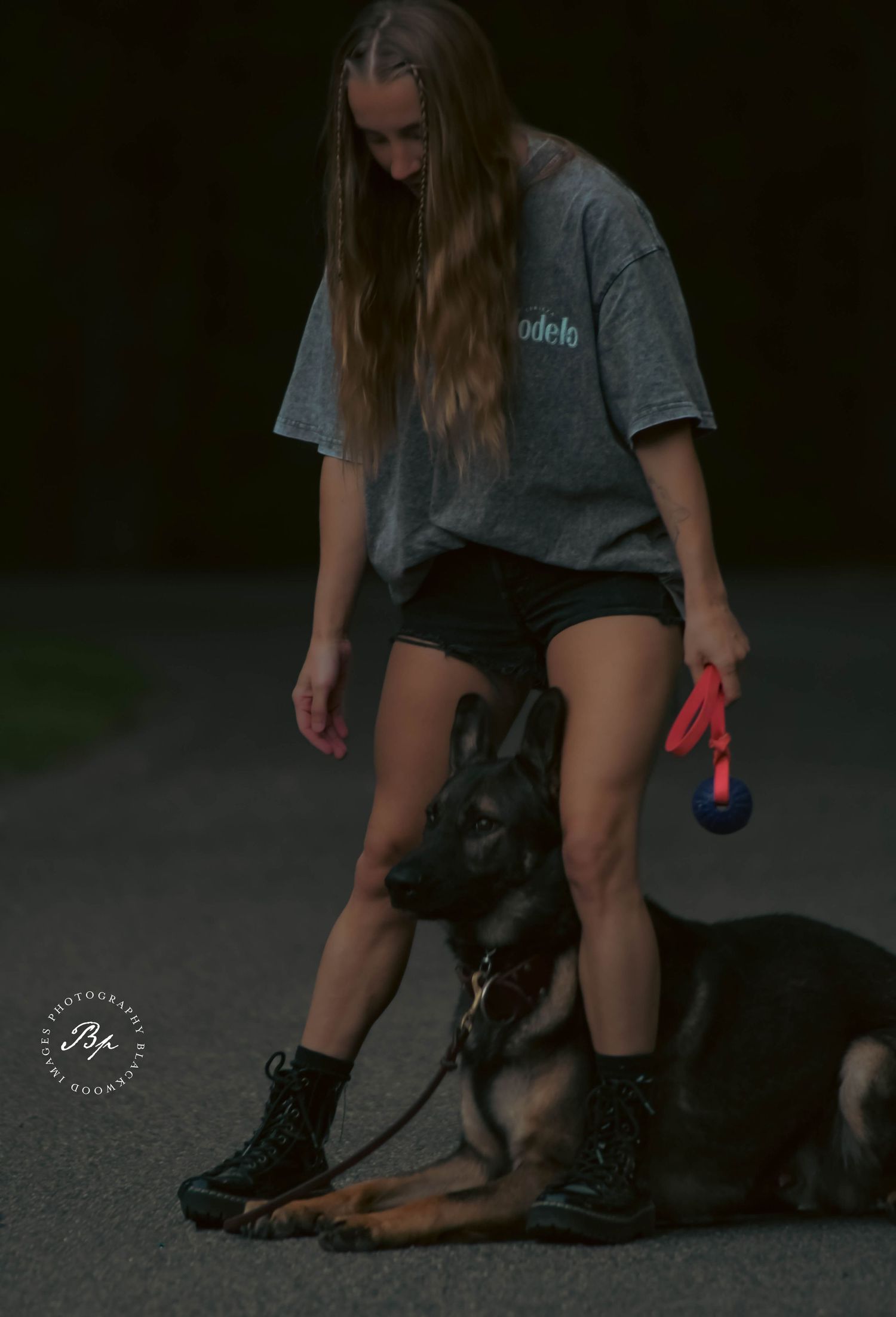 Young woman in gray Modelo shirt and black shorts walks a German Shepherd dog on a red leash at dusk.
