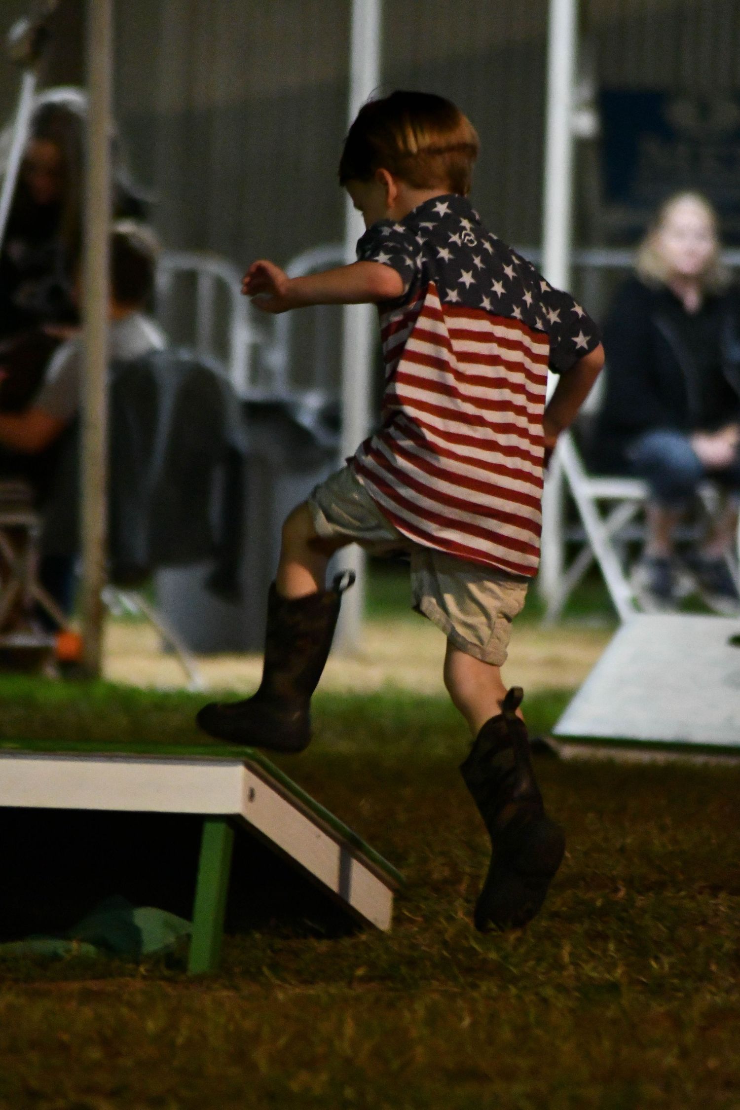 Young boy in American flag shirt and cowboy boots jumping on a cornhole board.