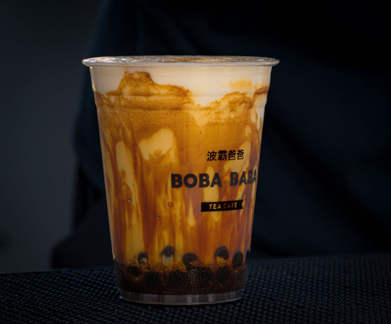 Boba Bar milk tea with caramel swirls in a clear plastic cup on a dark background.