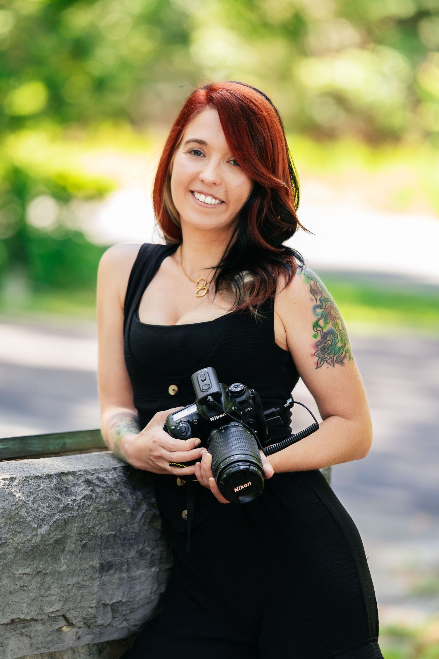 Smiling woman with red hair holding a Nikon camera outdoors, wearing black outfit with water color tattoo on shoulder. Branding Photo.