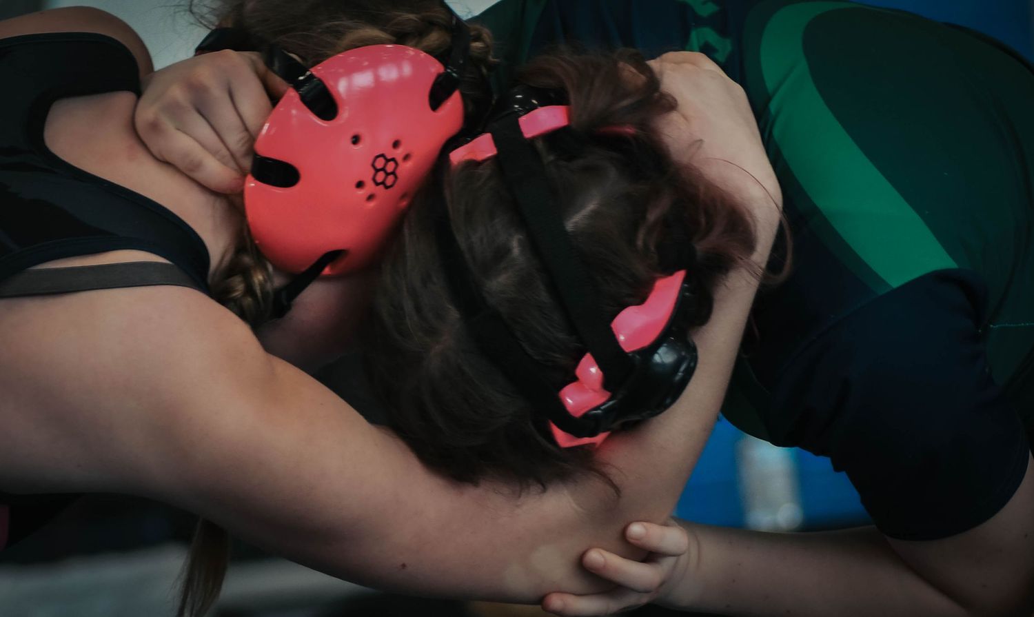 Two female wrestlers grappling on a mat, both wearing pink and red protective headgear.