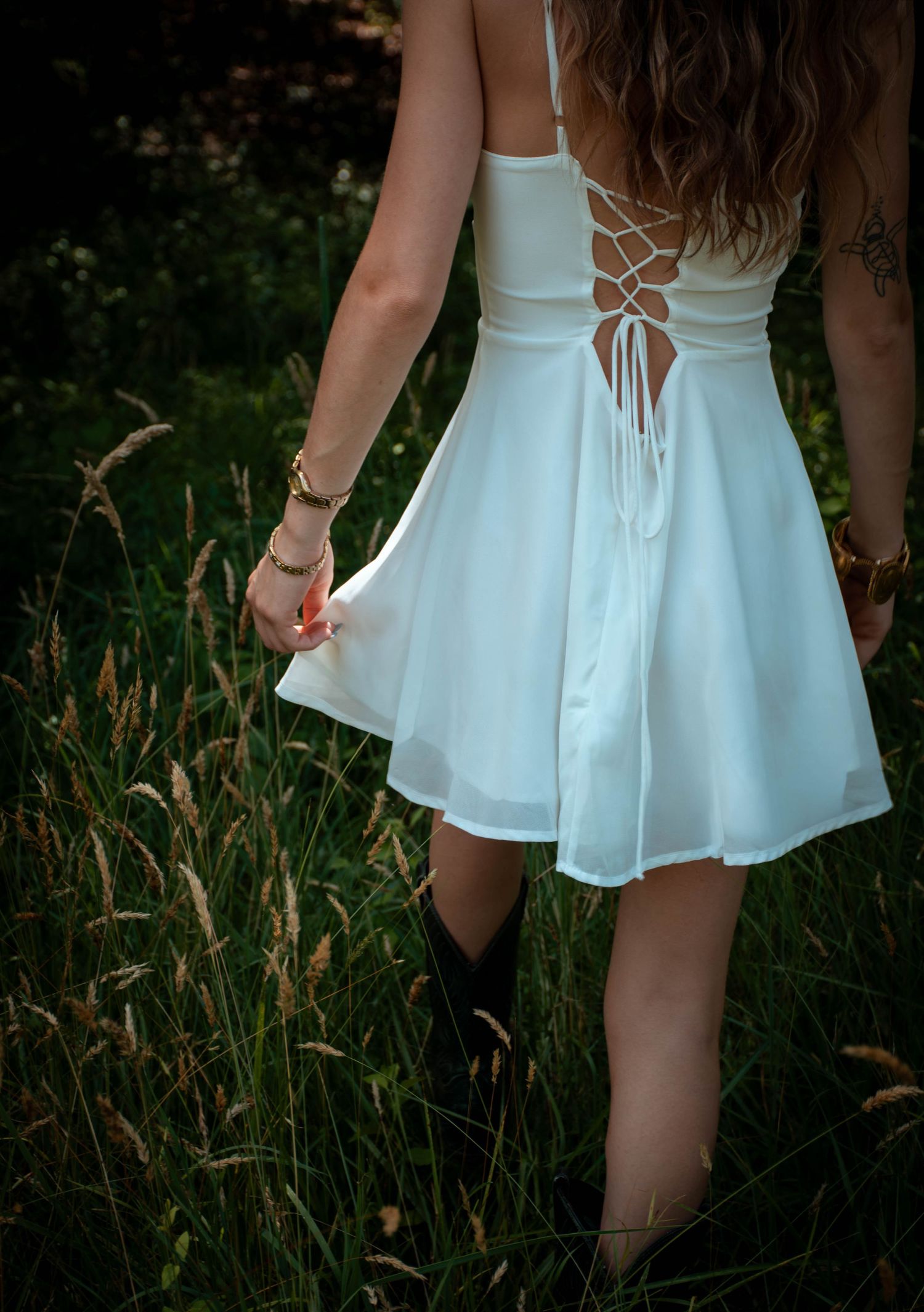 Woman in white lace-back dress and black cowboy boots walking through tall grass in a lush green field.