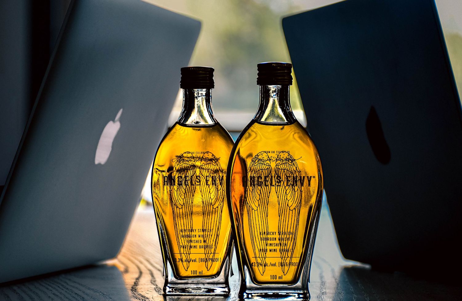 Two Angel's Envy bourbon miniature bottles on table between Apple laptops in sunlight.