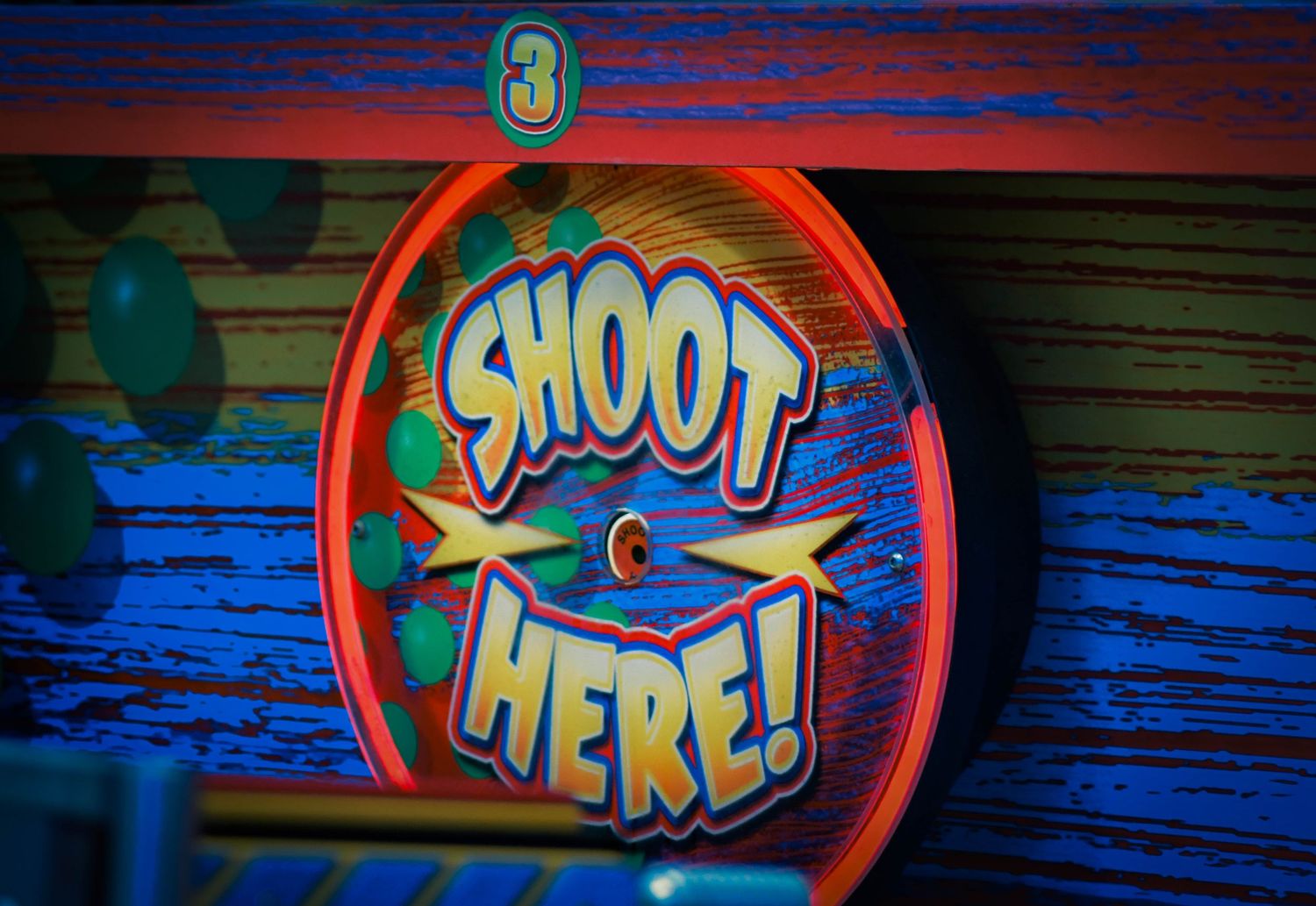 Colorful arcade game sign with neon lights reading Shoot Here, featuring star bursts and green dots on a wooden wall.