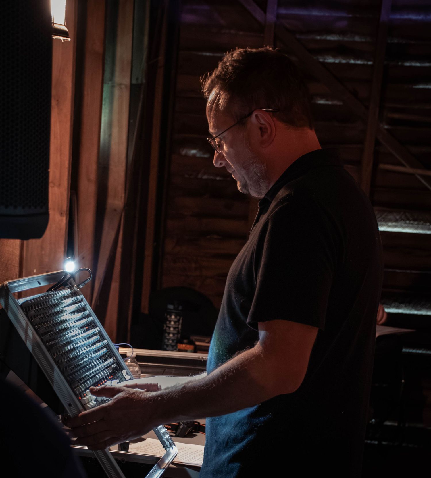 Man with glasses operating a mixing console in a dimly lit recording studio or live music venue.