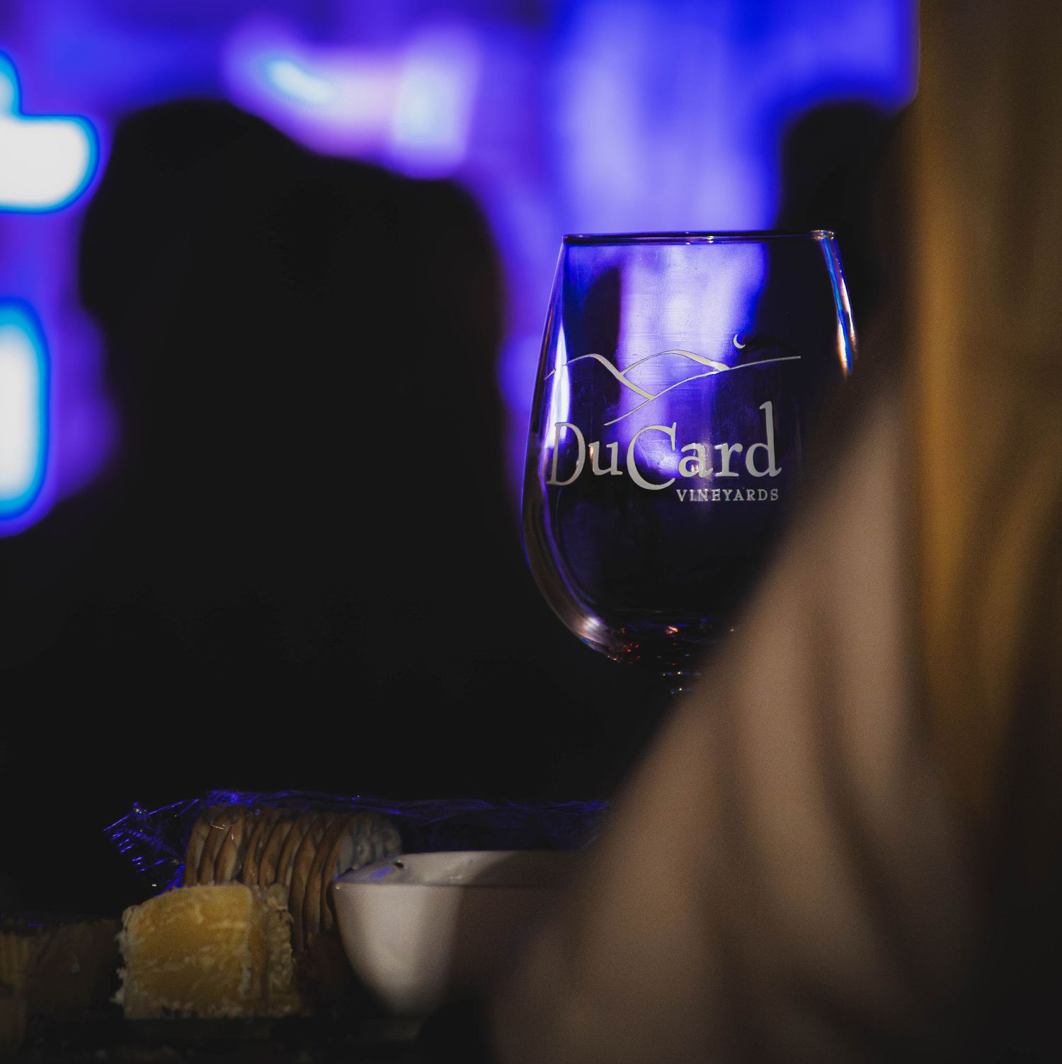 DuCard Vineyards branded wine glass on a dimly lit table with food and purple ambient lighting.