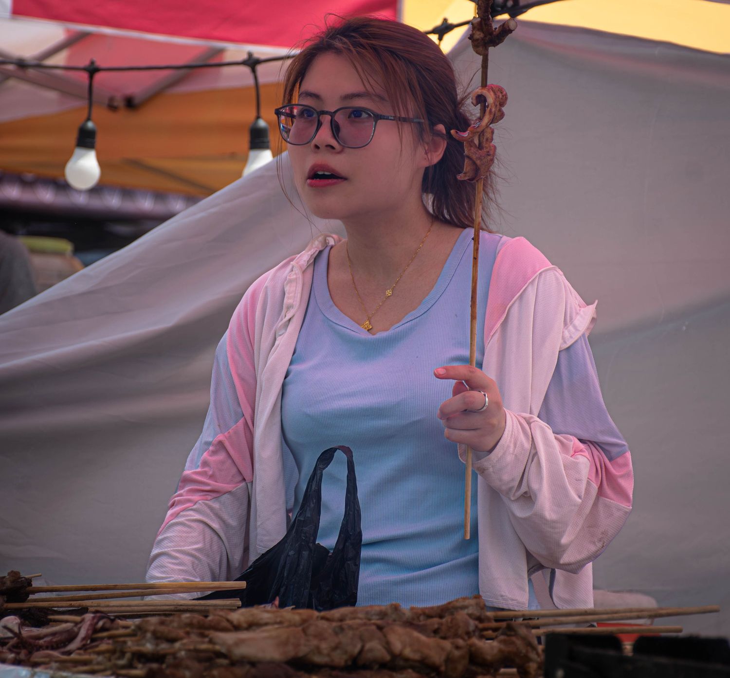 Young woman with glasses selling food at an outdoor market stall, wearing a pink jacket over a blue shirt.