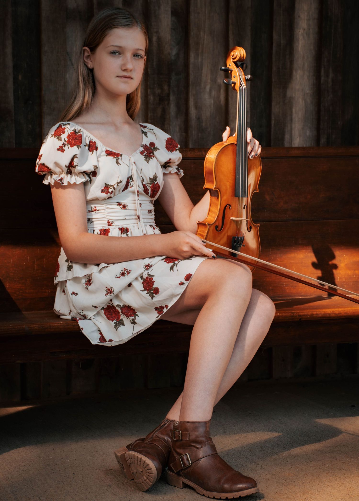 Young girl in floral dress holding a violin and bow, seated on a bench in warm golden light.