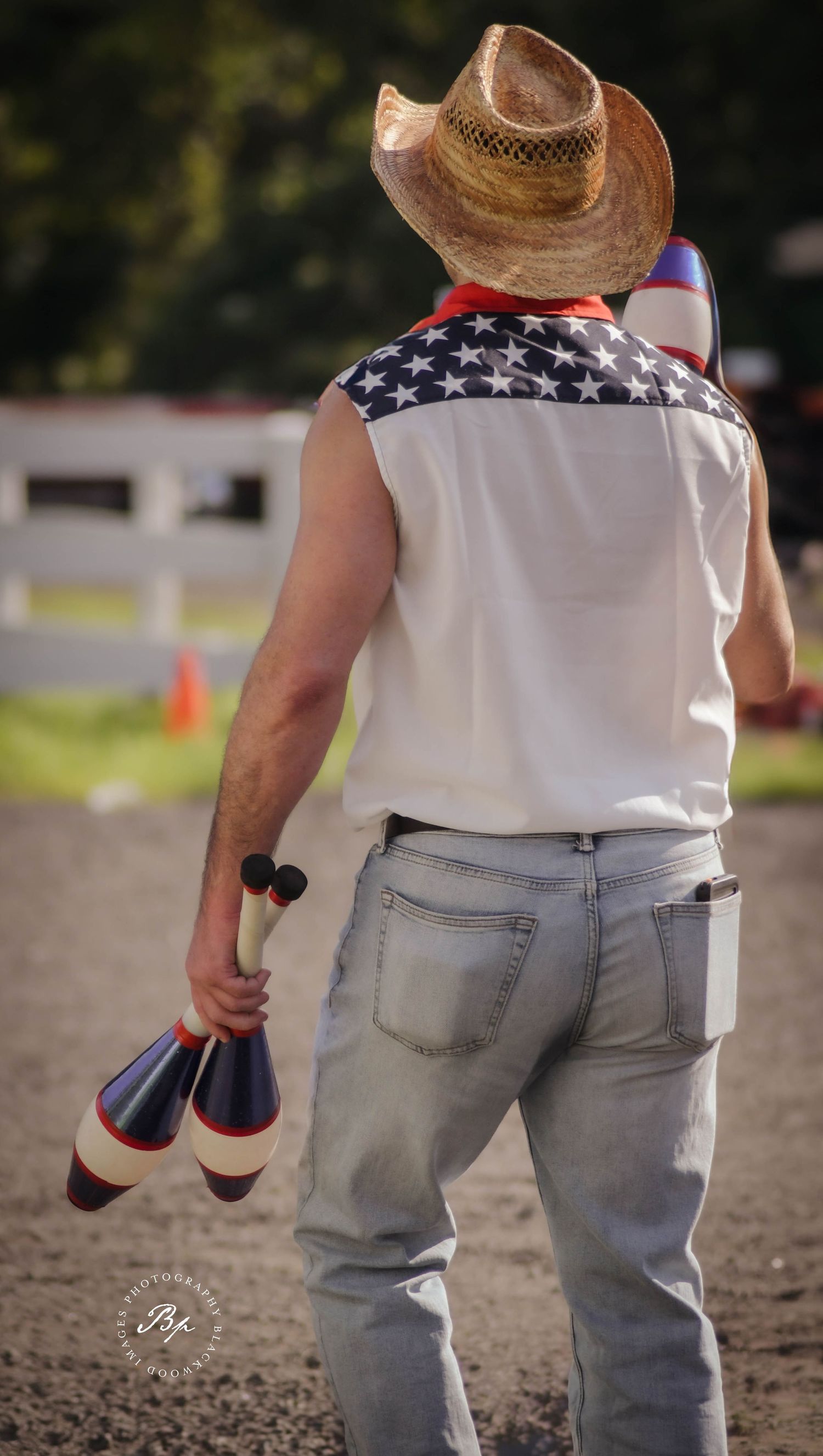 Man in American flag vest and cowboy hat holding juggling pins at outdoor event.