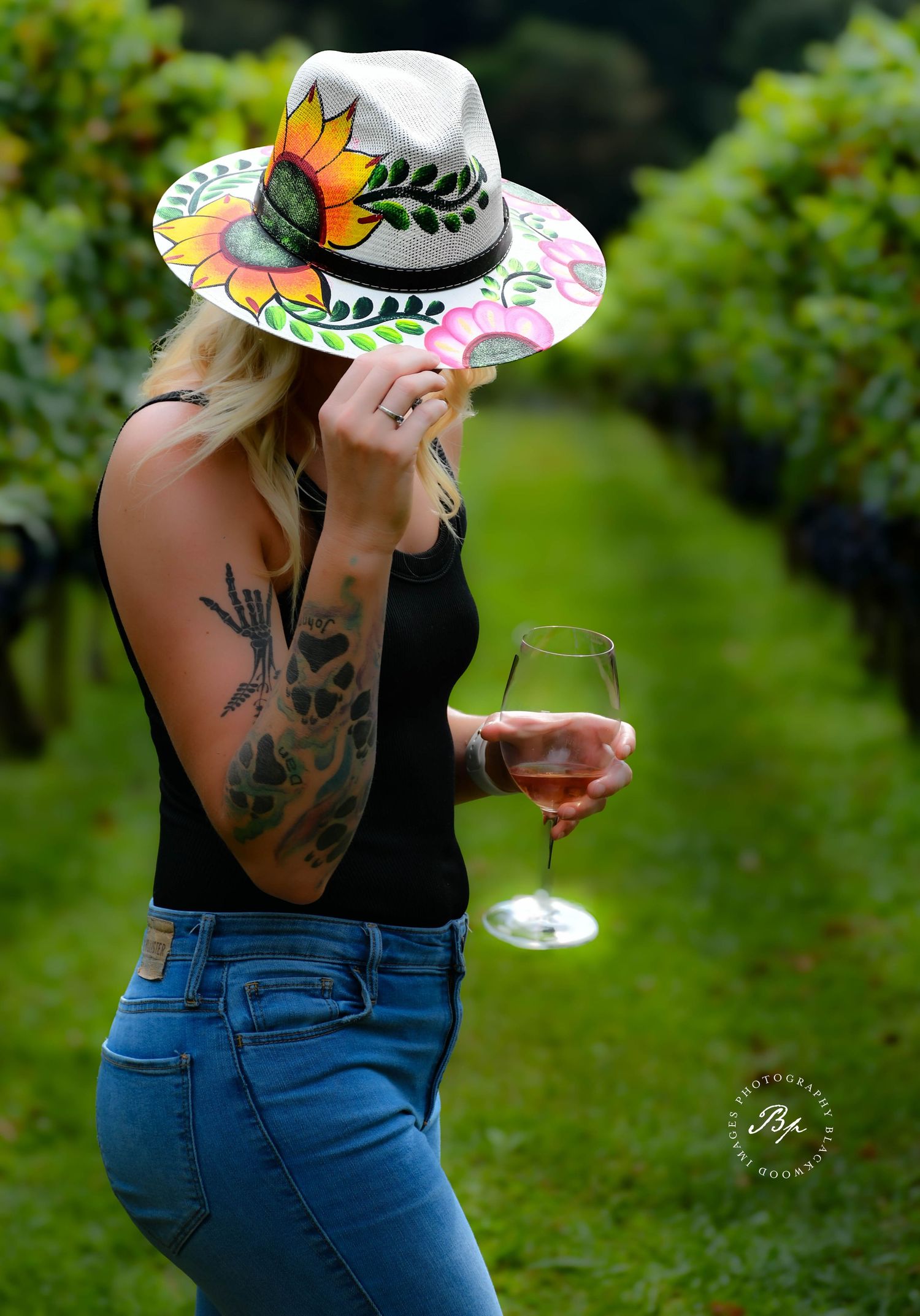 Tattooed blonde woman holding wine glass wearing colorful painted sunflower hat in vineyard.