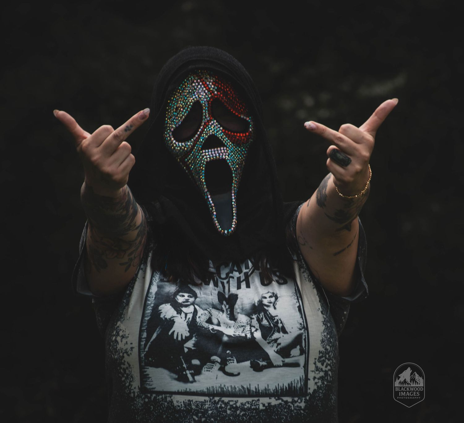 Person wearing a bejeweled Ghostface mask and horror graphic tee, flashing rock on hand signs outdoors.