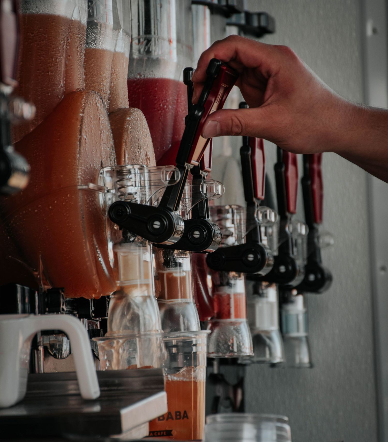 Hand operating a tap on a row of colorful slushie drink dispensers filled with pink and orange frozen beverages.