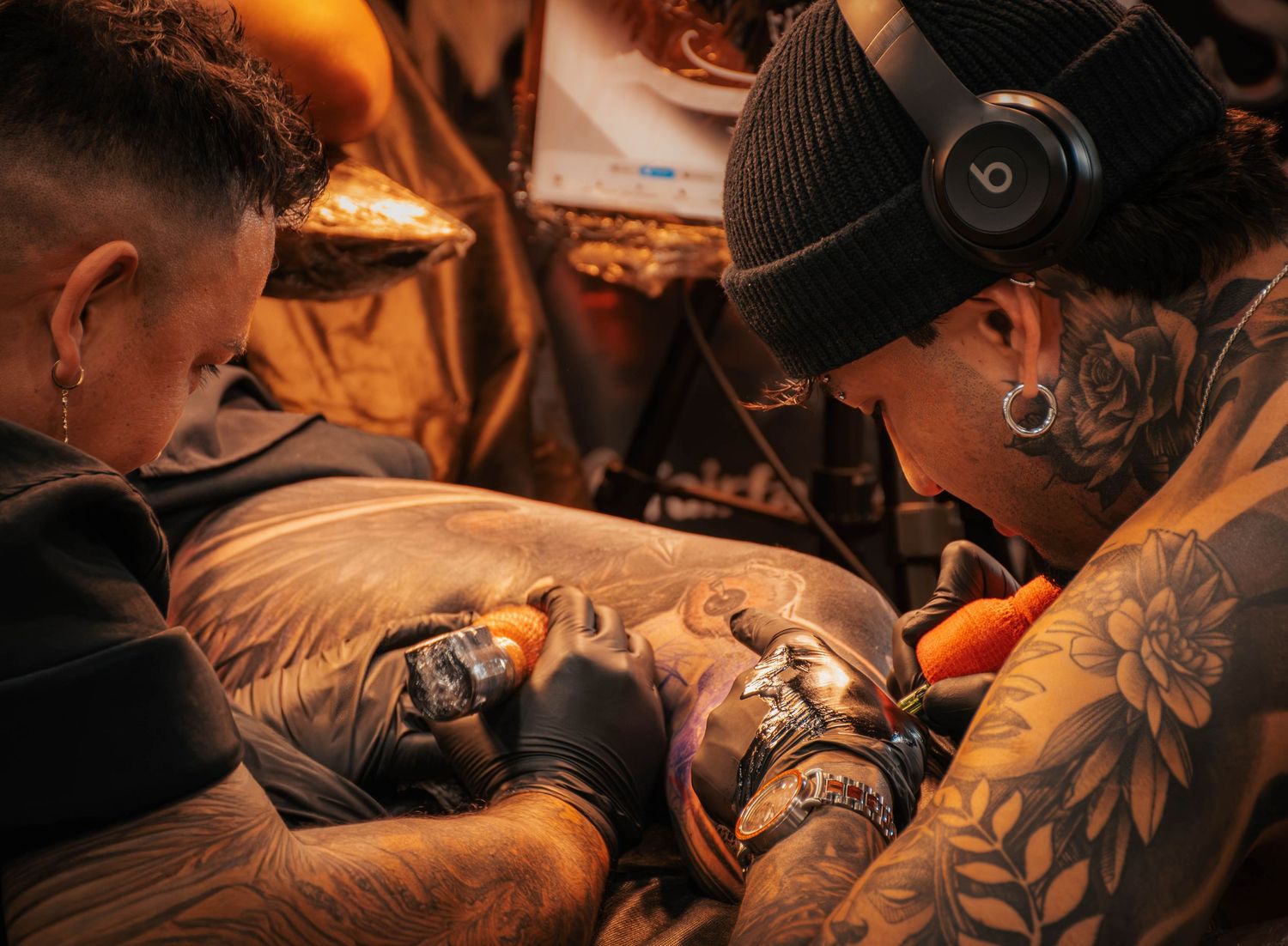 Two tattoo artists with tattoos and gauges collaborating on a detailed tattoo design in a studio.