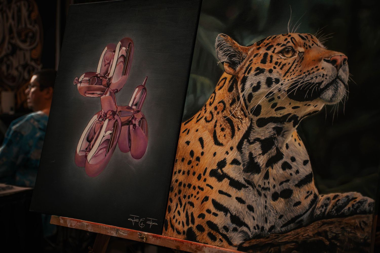 A jaguar rests beside an easel displaying a pink balloon dog artwork in a dimly lit, ornate setting.
