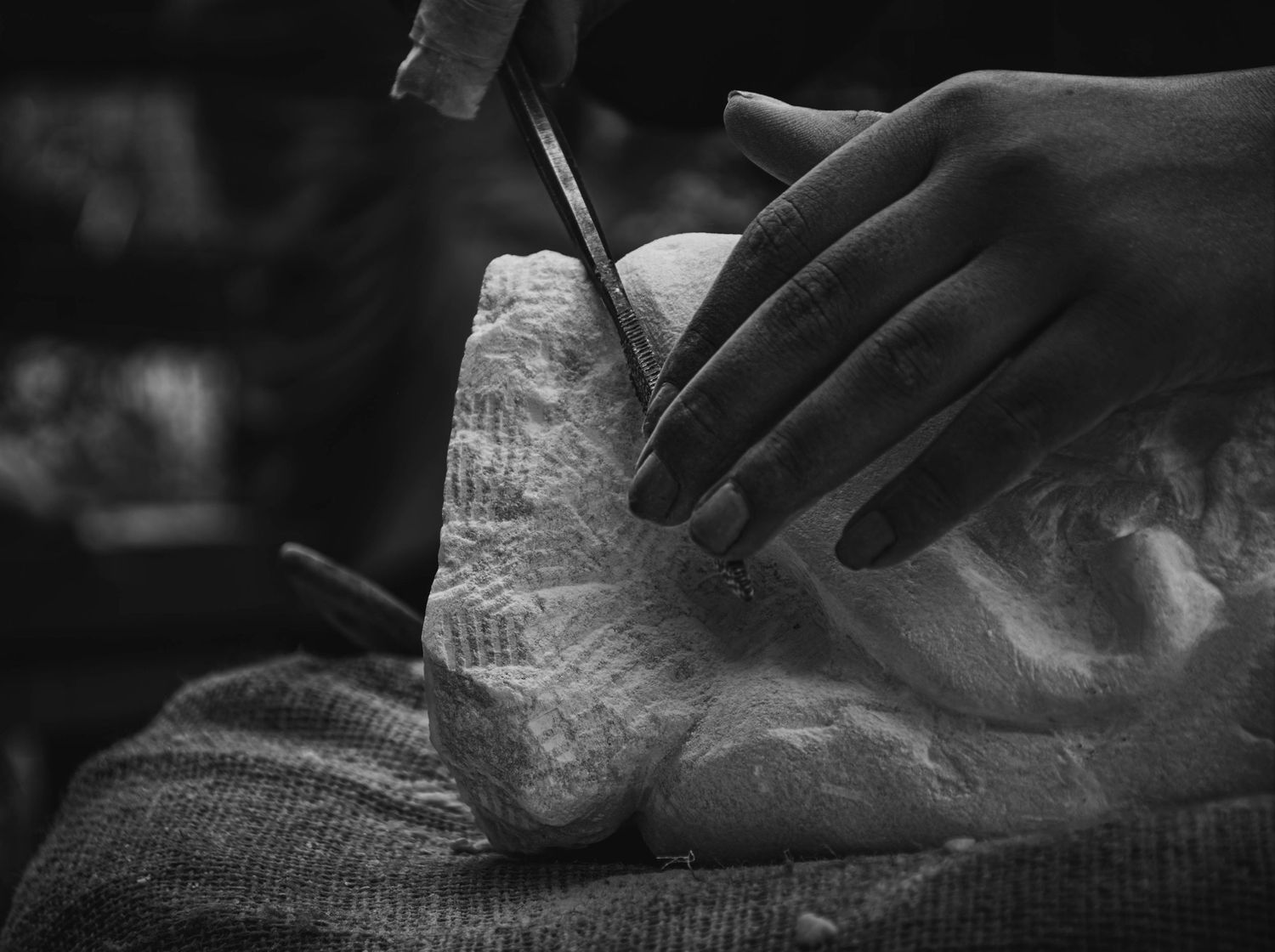 Hands carefully carving a white stone sculpture using a chisel tool in a black and white photograph.