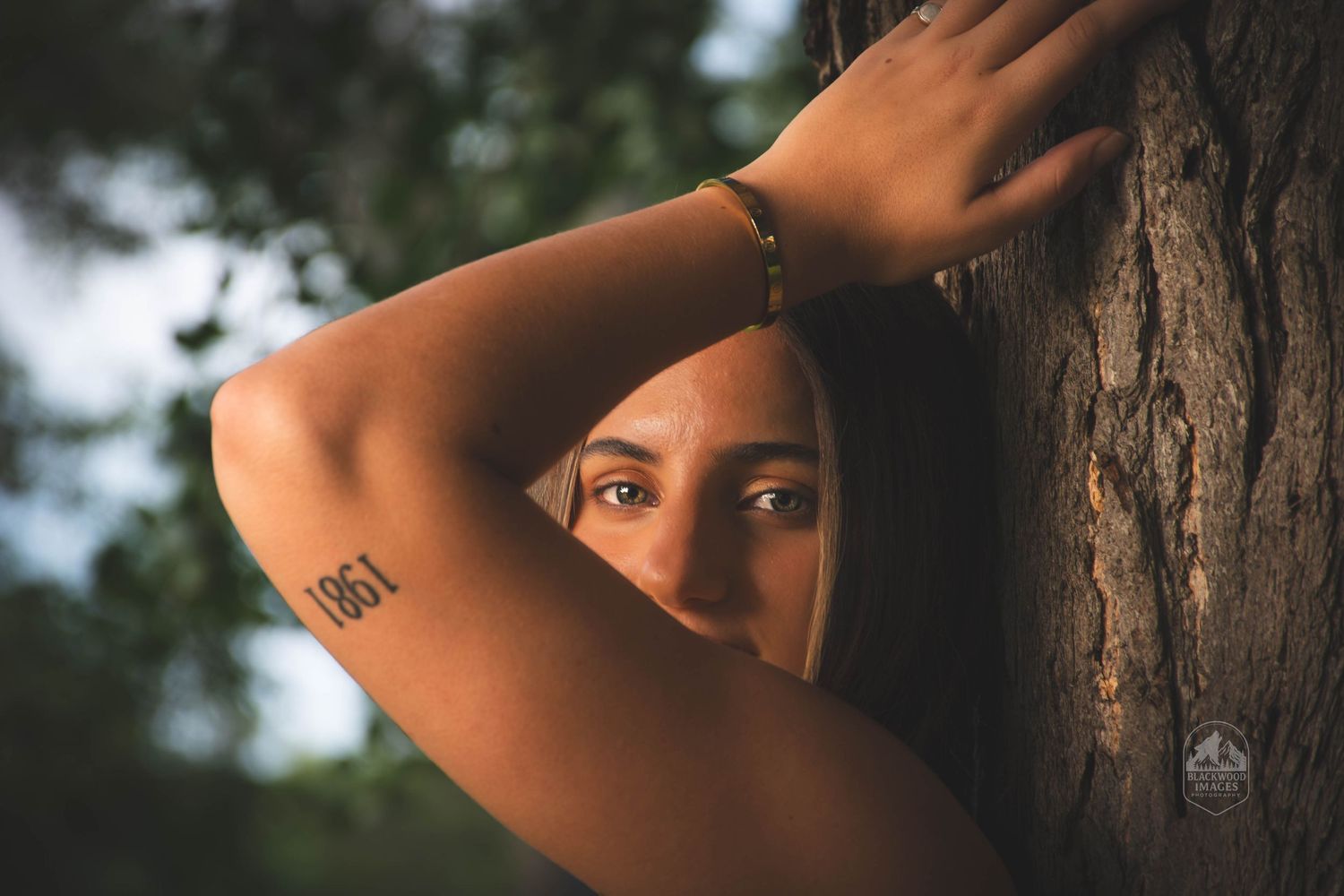 A young woman with a '1861' tattoo on her arm peeks around a tree trunk outdoors.