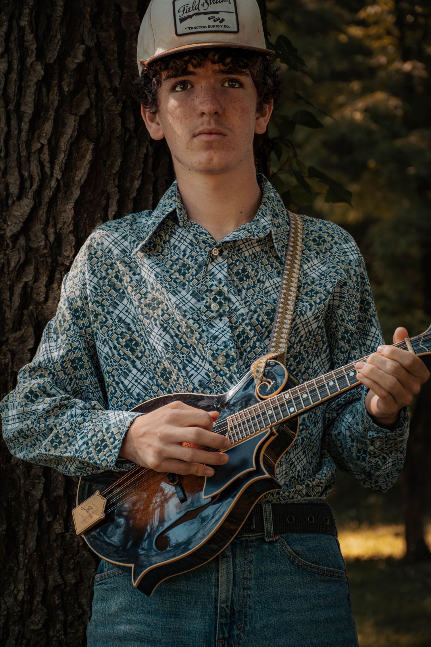Young musician playing mandolin outdoors, wearing floral shirt and trucker hat, leaning against tree in natural light.