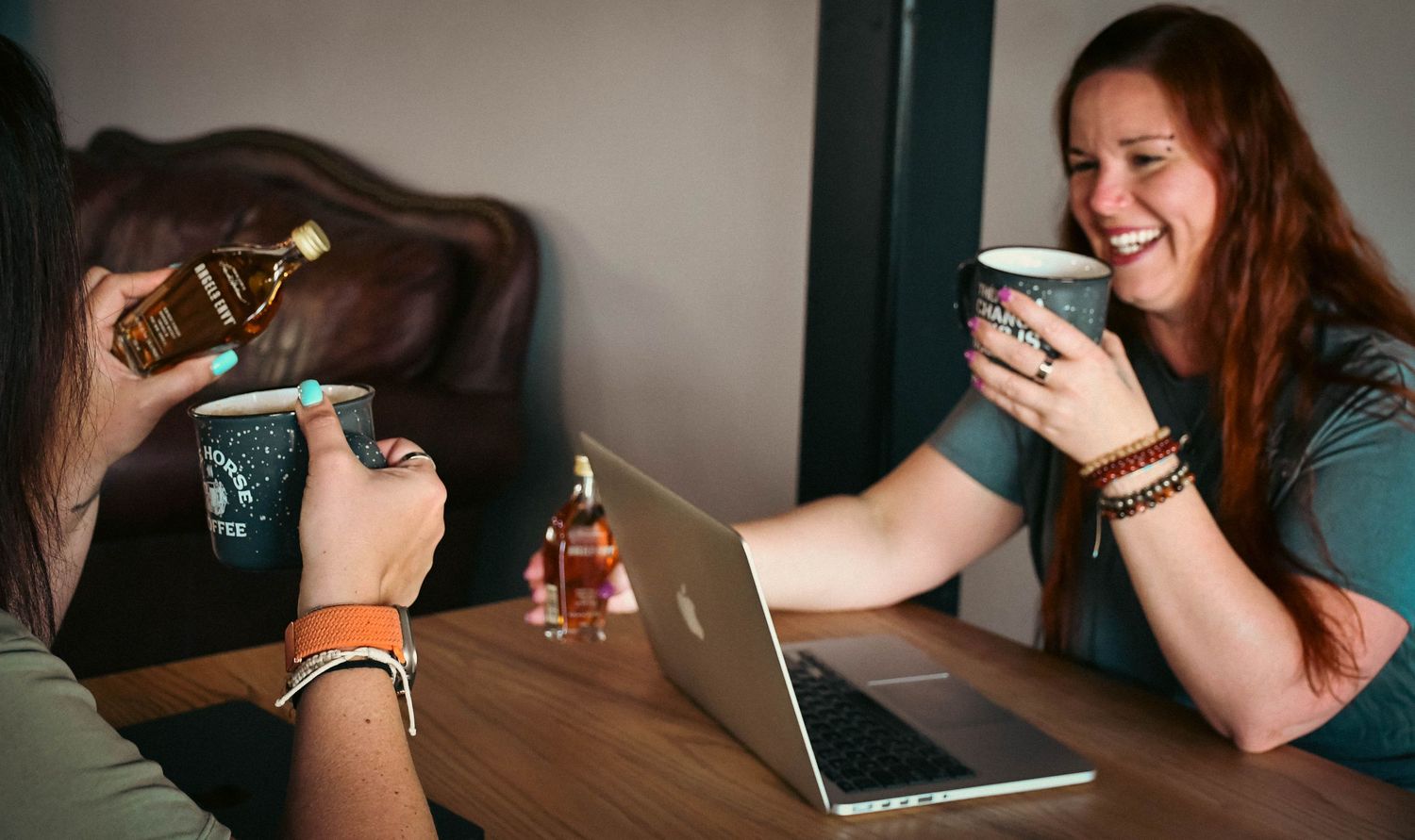 Two women laughing and toasting with coffee mugs and bourbon bottles on a wooden table.