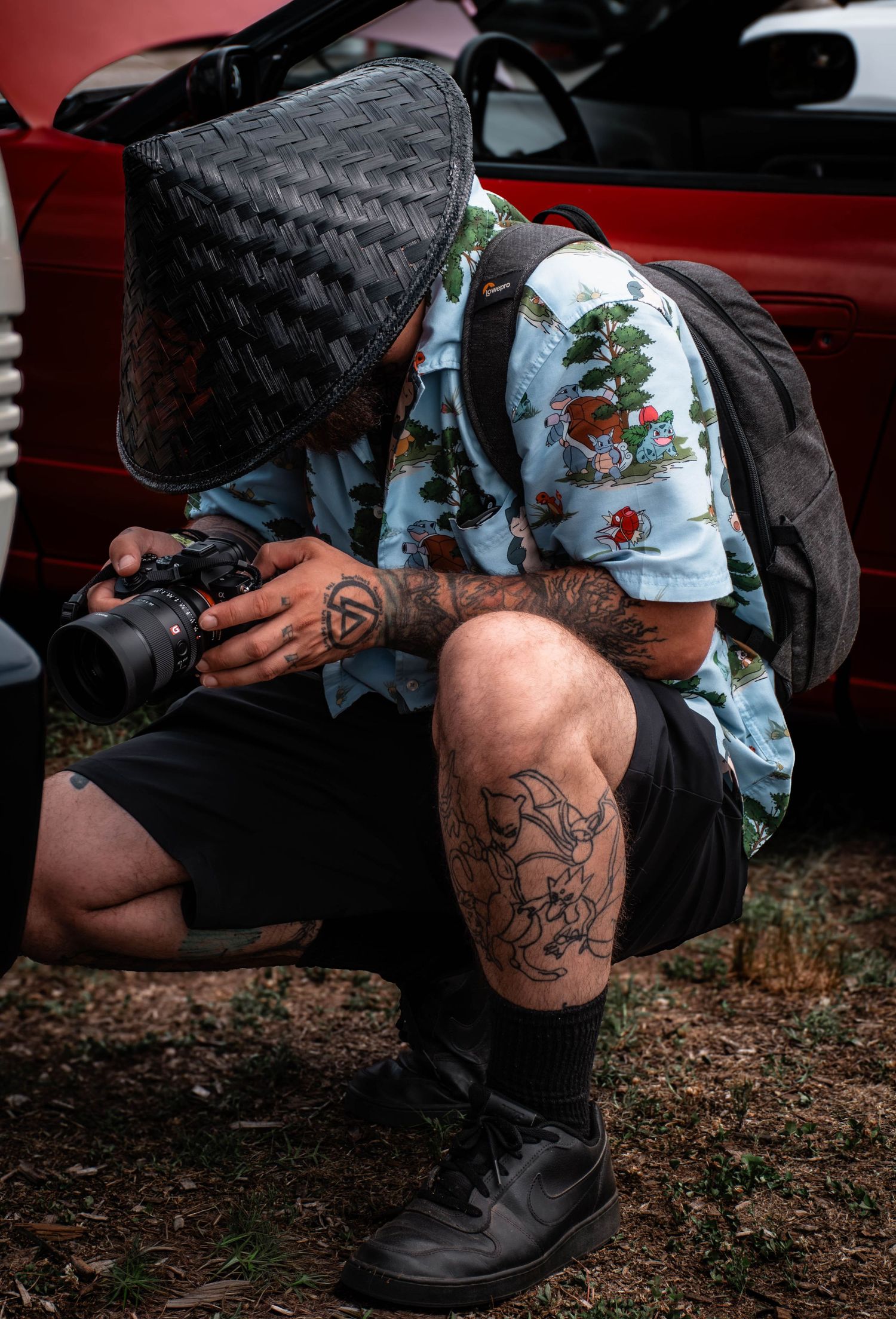 Tattooed photographer crouching in Pokemon Hawaiian shirt and woven hat, reviewing shots on camera near a red car outdoors.