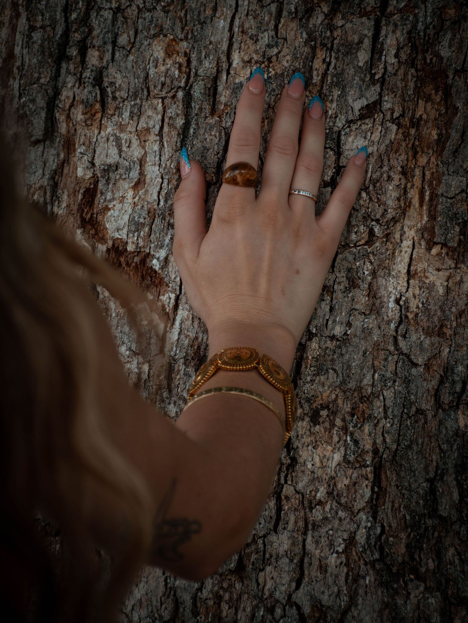 A hand with teal nails, gold bracelet, and rings rests on rough tree bark, with a small wrist tattoo visible.