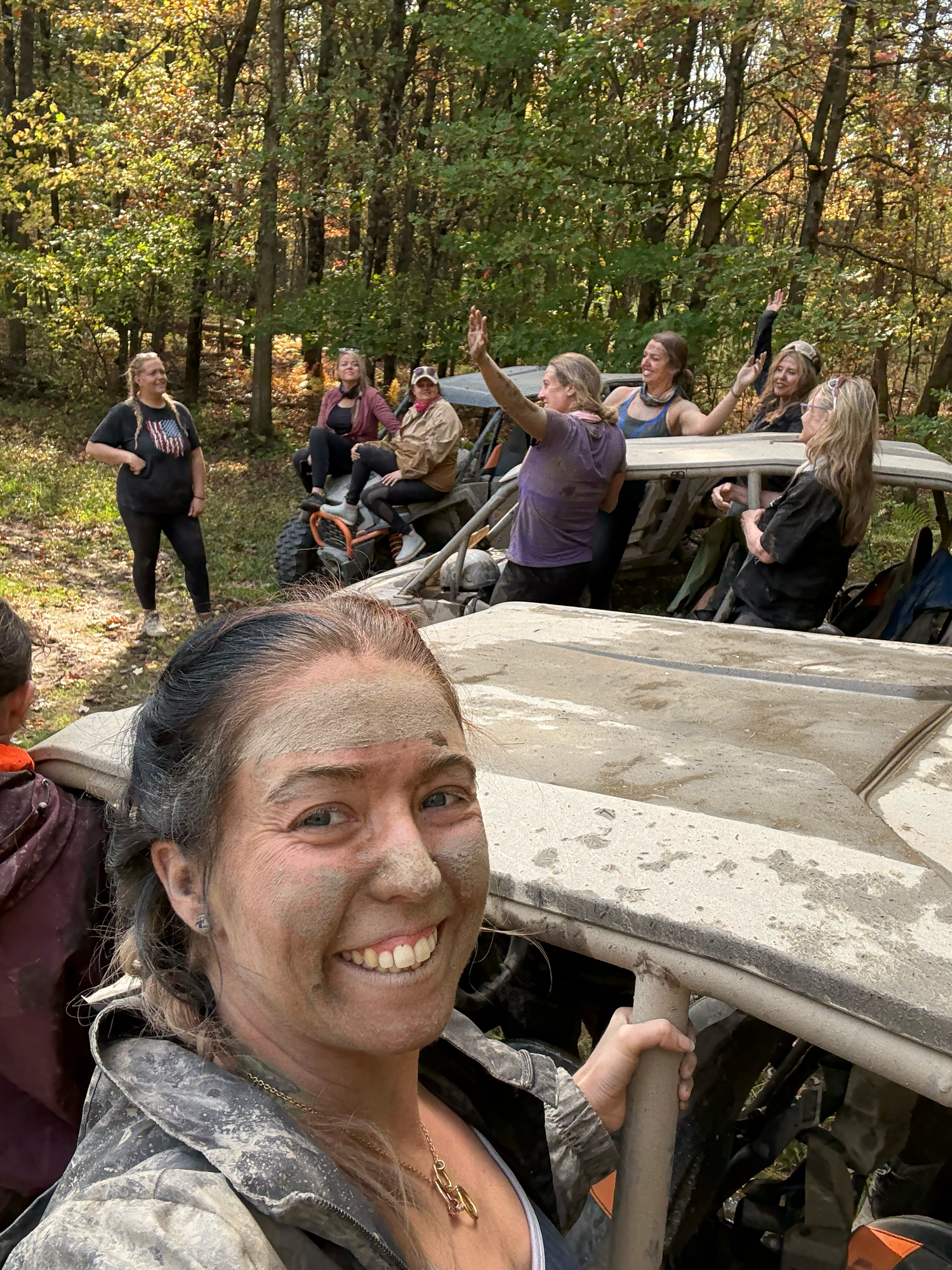 Woman with mud on her face smiling during an outdoor ATV trail ride with a group of friends in autumn woods.