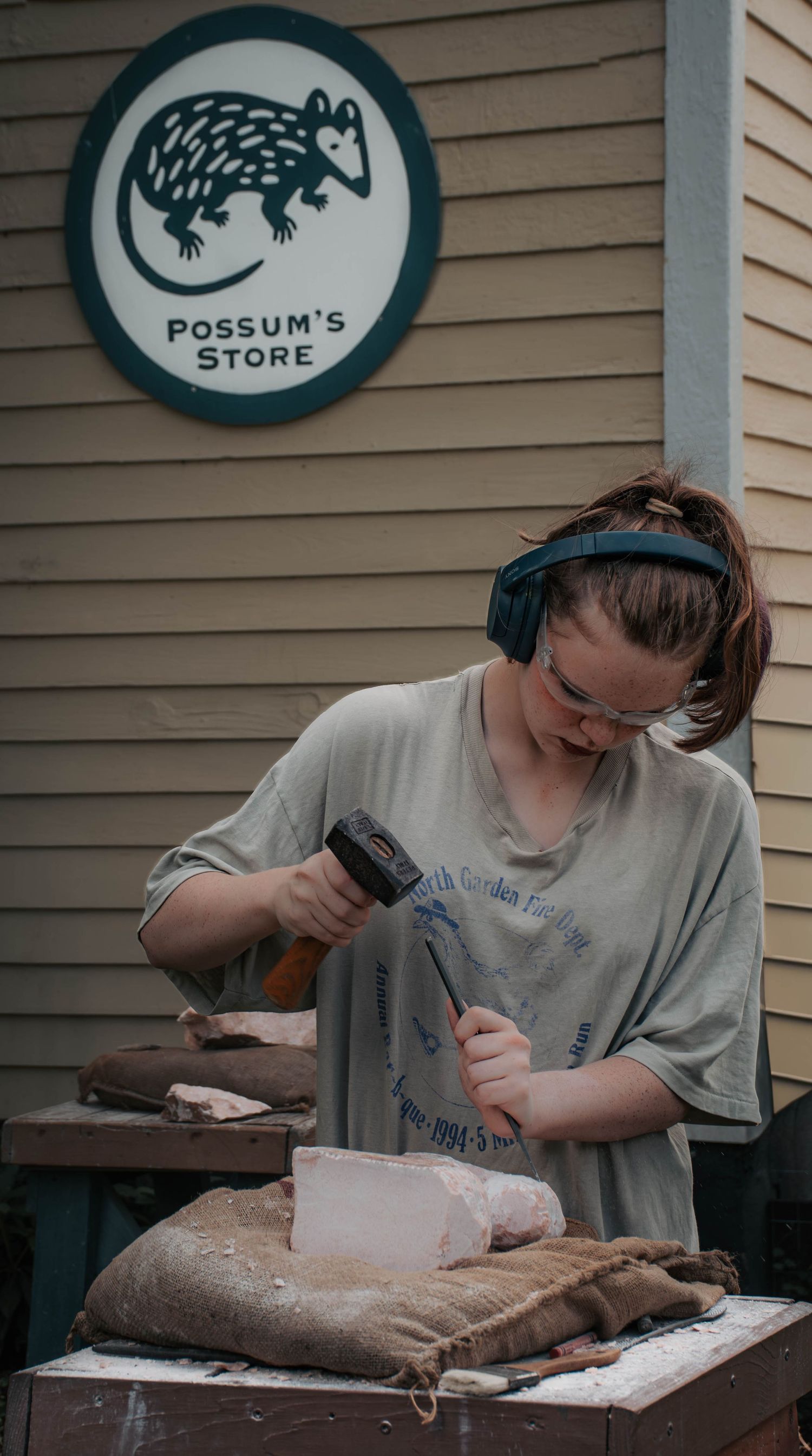 Young woman carving stone with hammer and chisel outside Possum's Store wearing safety gear.