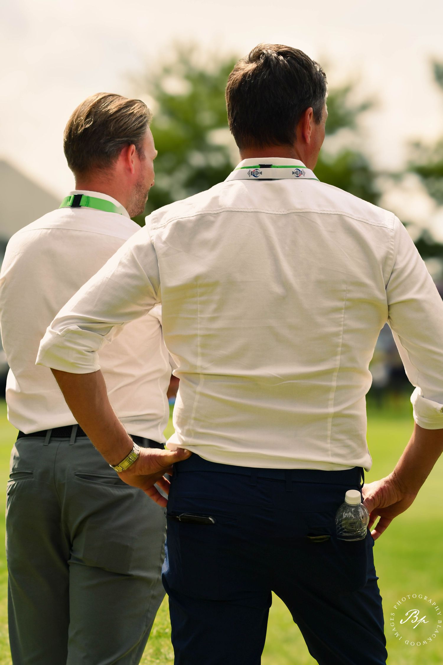 Two men in white dress shirts viewed from behind at outdoor golf event.