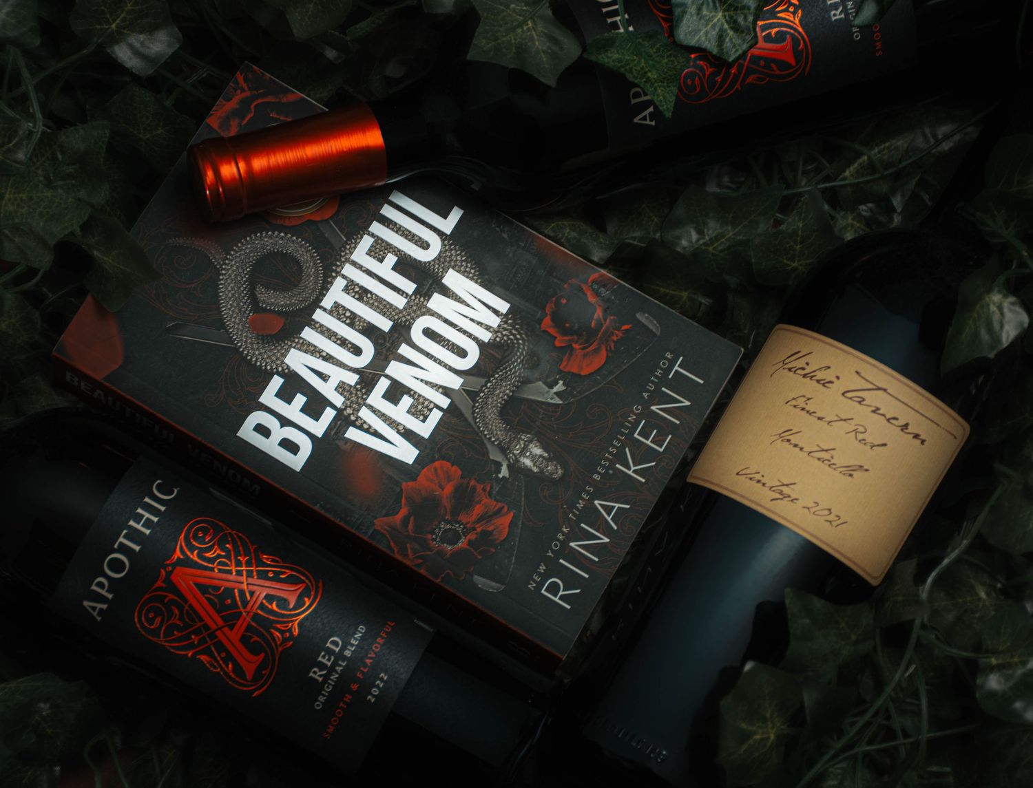 Book 'Beautiful Venom' by Rina Kent surrounded by dark wine bottles including Apothic, on lush green ivy background.