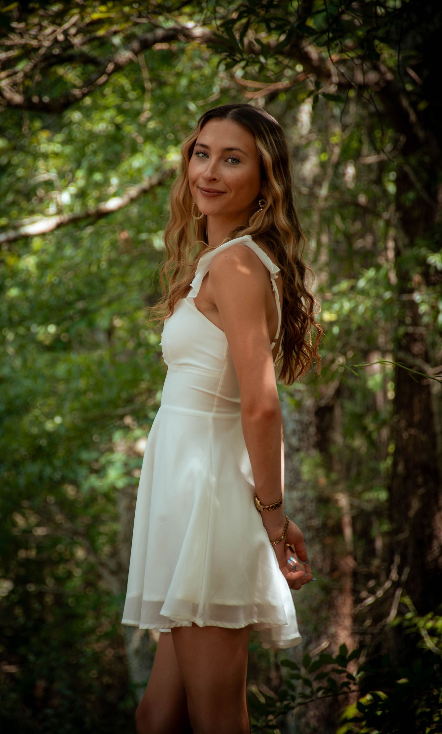 Young woman in white mini dress posing gracefully among lush green trees in a sunlit forest setting.