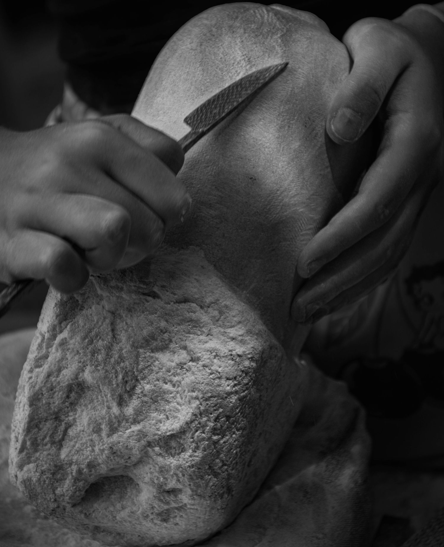Hands sculpting a stone with a chisel tool in a black and white close-up photograph.