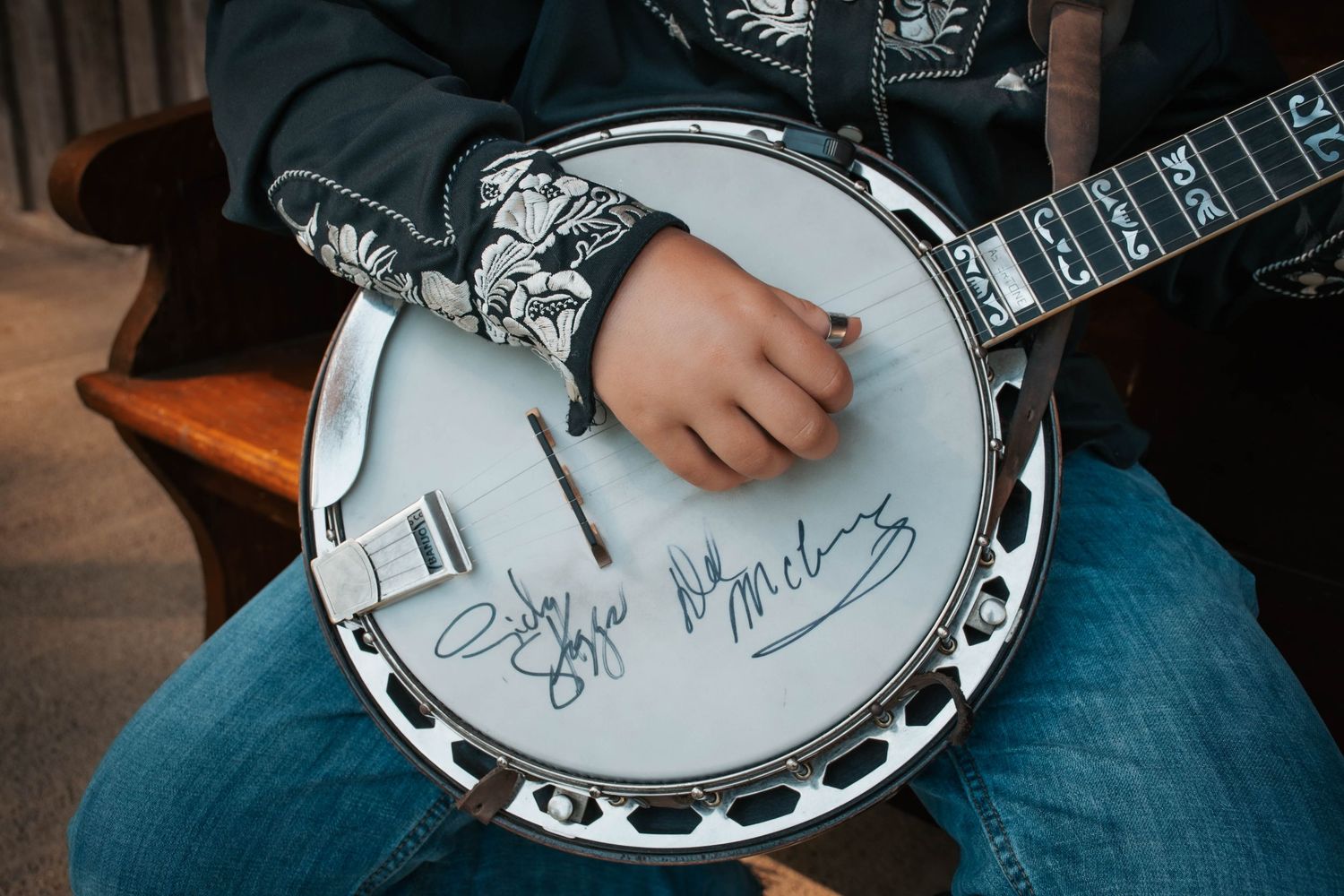 Close-up of hands playing a signed banjo with autographs on the drum head, worn by a musician in a dark jacket.