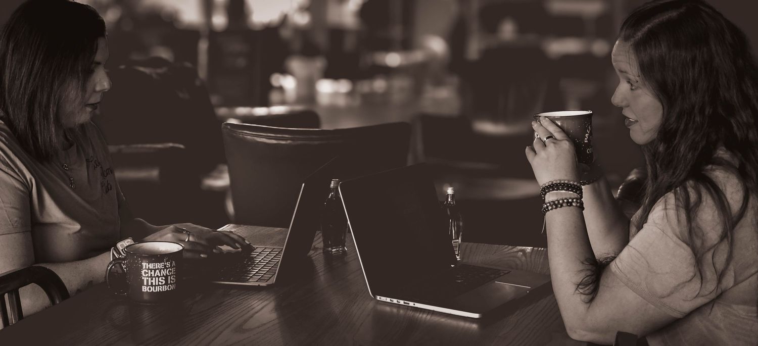 Two women sit across from each other at a cafe table with laptops and coffee cups.