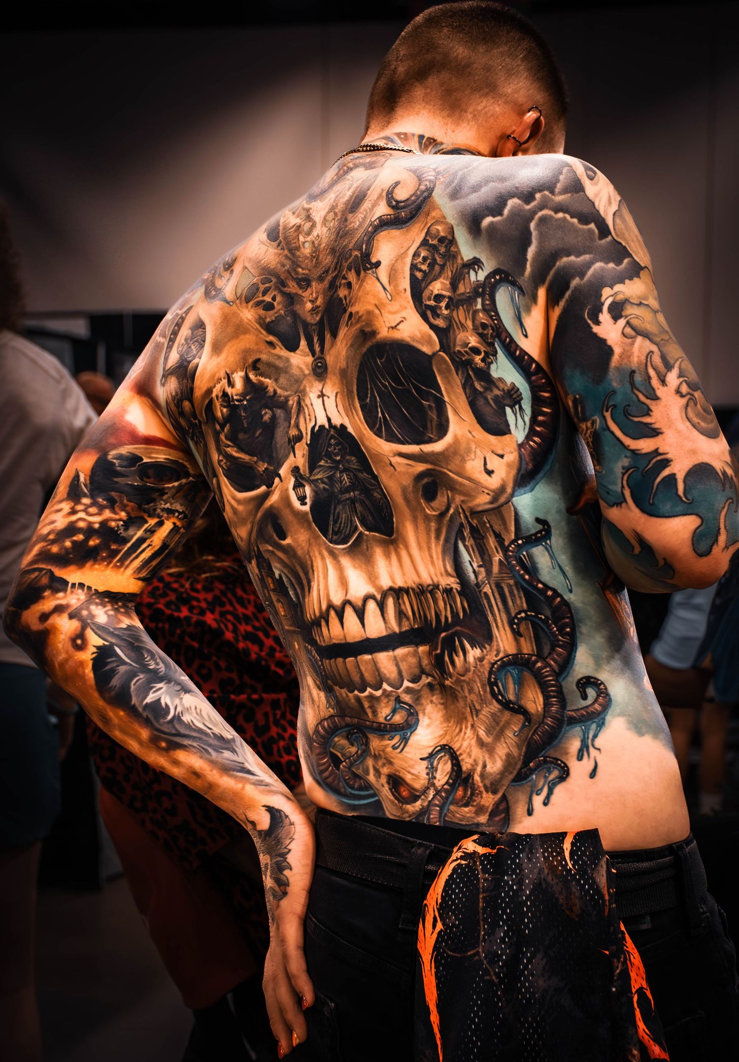 Man with full back tattoo featuring a large skull design with intricate black and grey detailing at a tattoo convention.