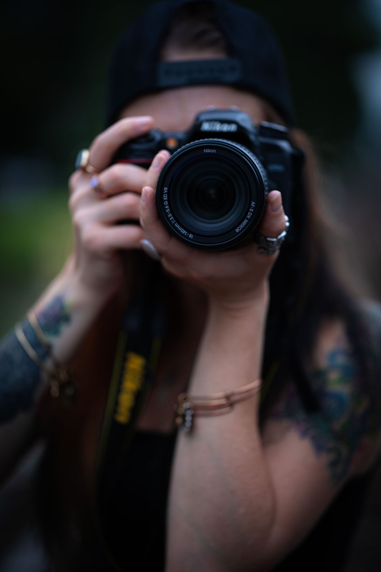 Woman with tattoos holding a Nikon DSLR camera up to her face, wearing a black cap outdoors.
