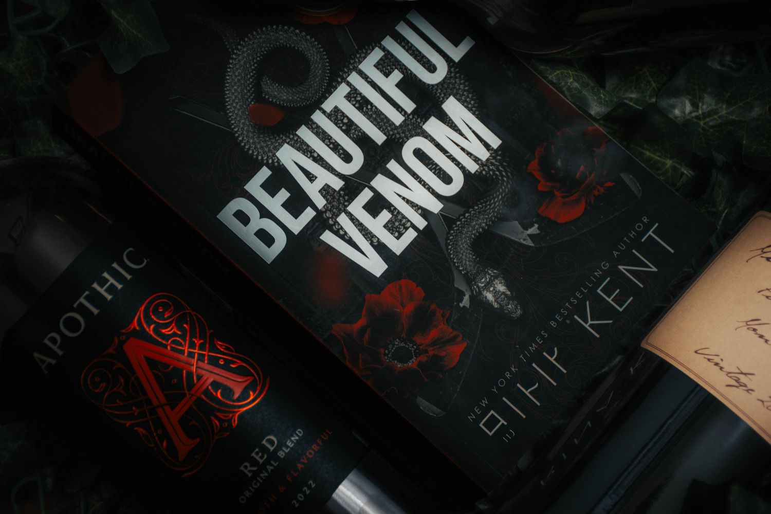 Dark styled flat lay featuring Beautiful Venom book beside Apothic Red wine bottle on dark background.