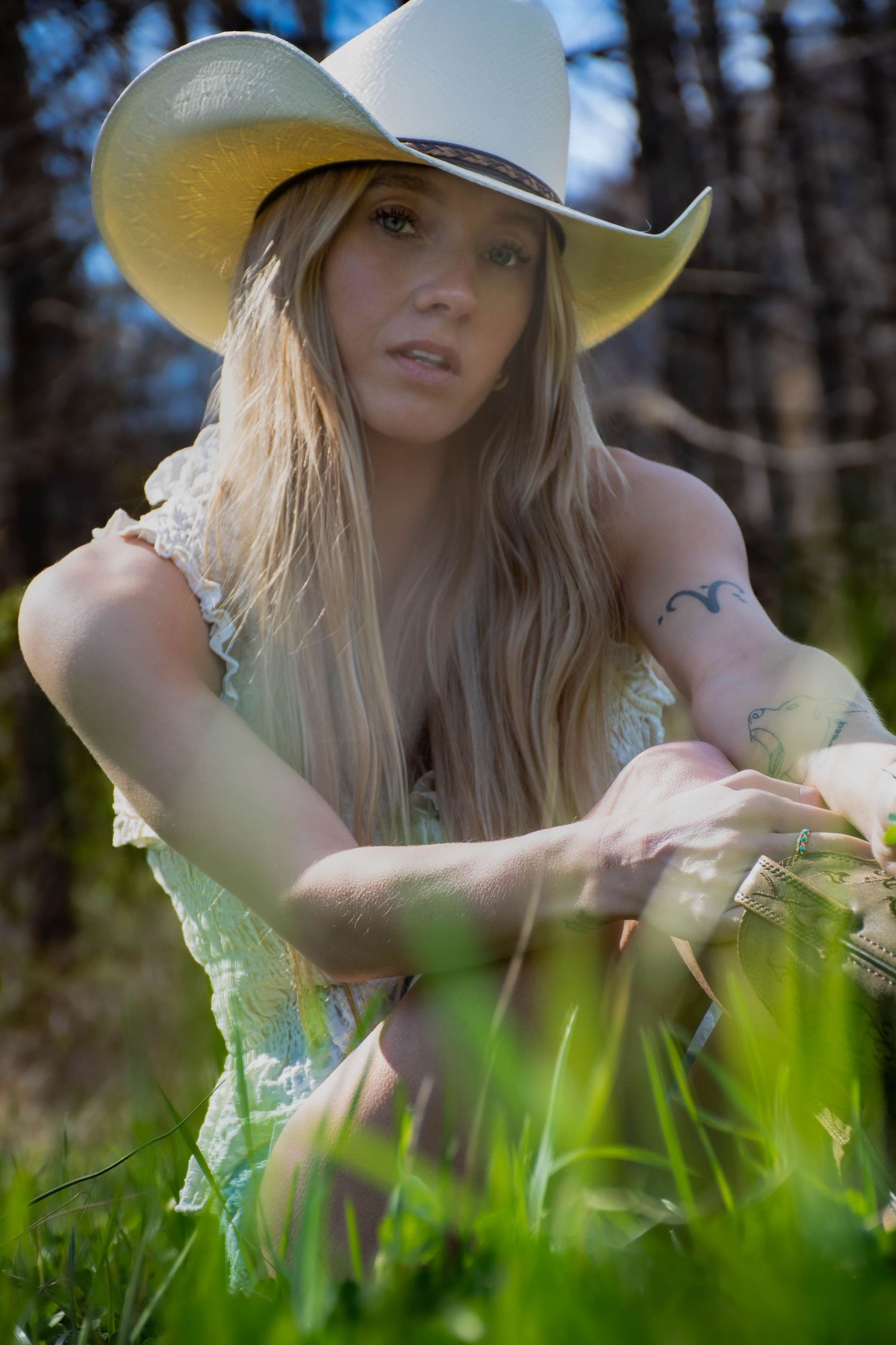 Blonde woman in white cowboy hat and lace dress poses in grass among trees.